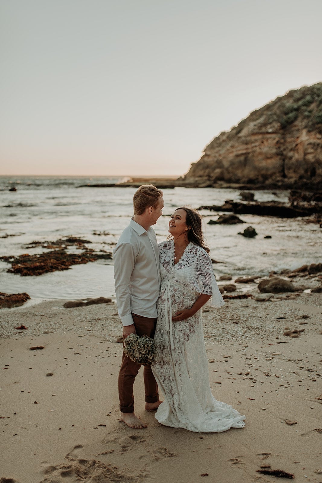 A couple standing on the beach during their elopement wedding. Bride is wearing a gorgeous lace dress from Everything Lace Hire  