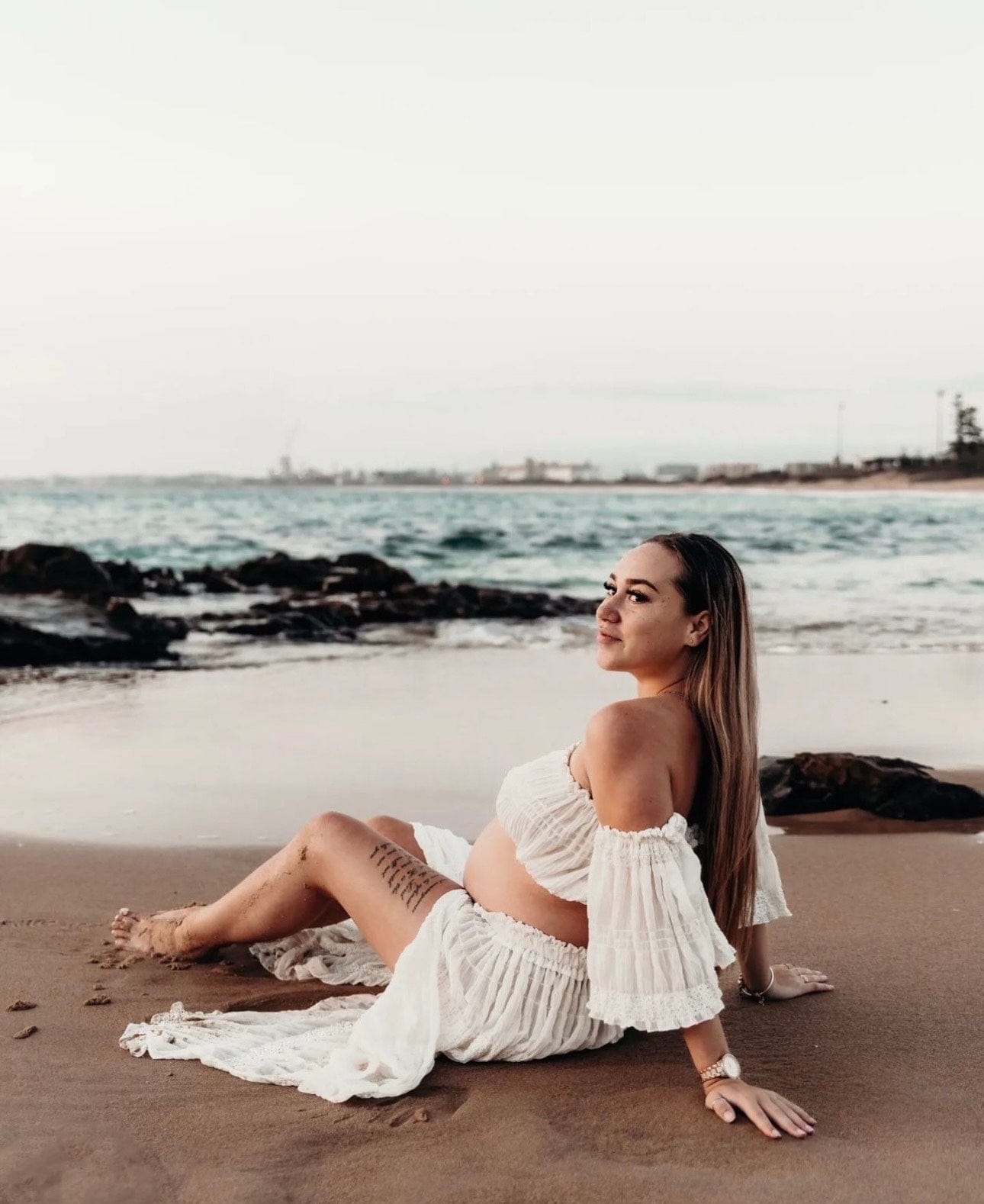 A pregnant lady sits on the sand as the waves come in wearing Everything Lace Hire Anabelle Two Piece Maternity Photoshoot Set