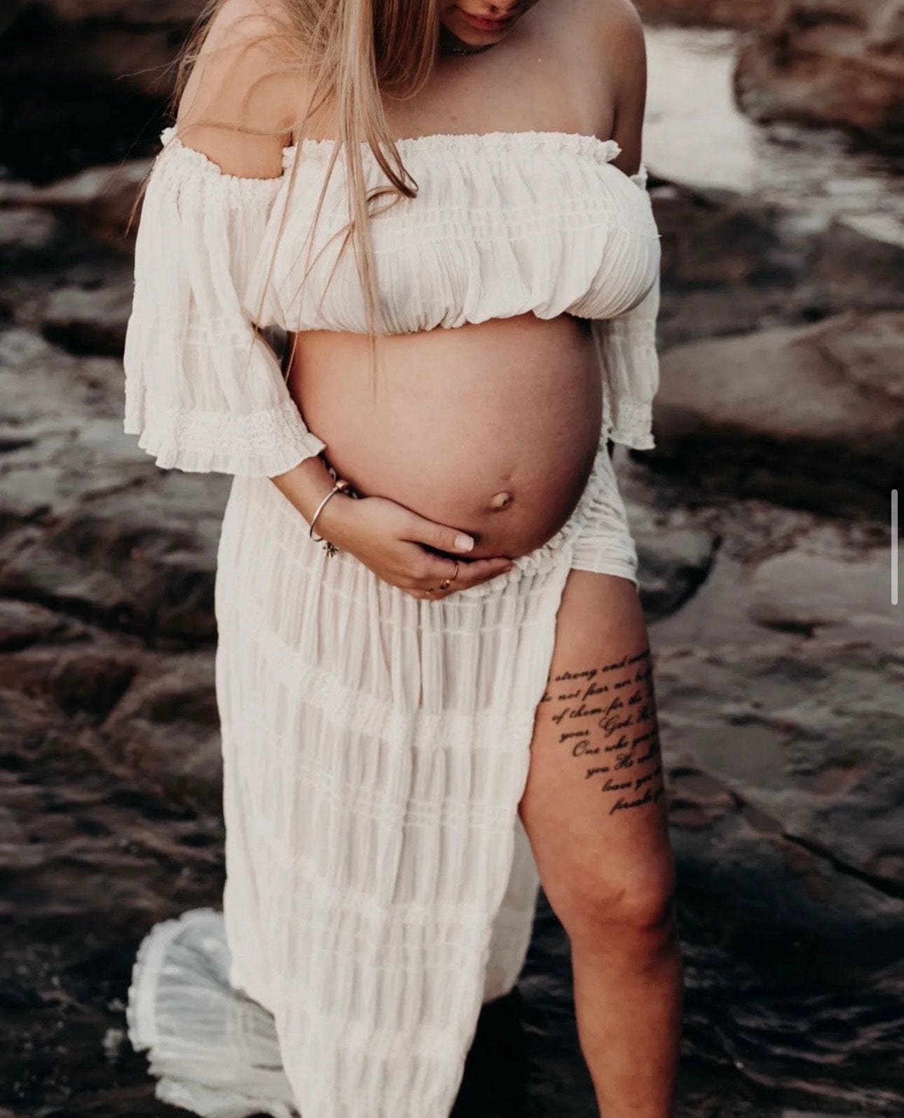 A pregnant lady stands amongst the rocks in the ocean holding her bump wearing Everything Lace Hire Anabelle Two Piece Maternity Photoshoot Set