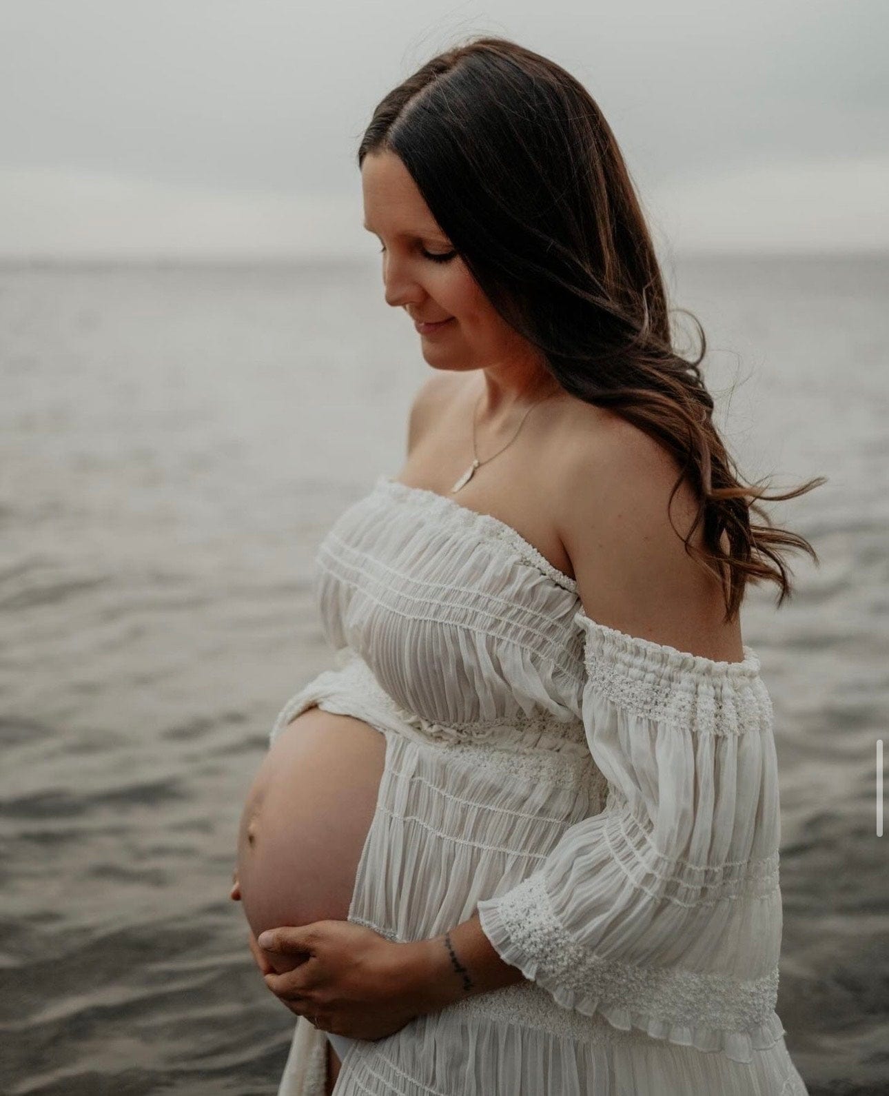 A pregnant lady stands in the sea looking at her pregnancy bump wearing Everything Lace Hire Anabelle Two Piece Maternity Photoshoot Set