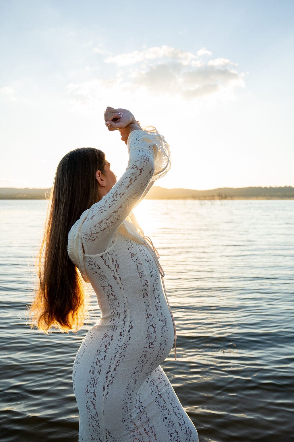 Pregnant lady wearing Everything Lace Hire House of CB Belle Dress a white lace dress standing by a lake with the sun setting behind her during her maternity photoshoot.