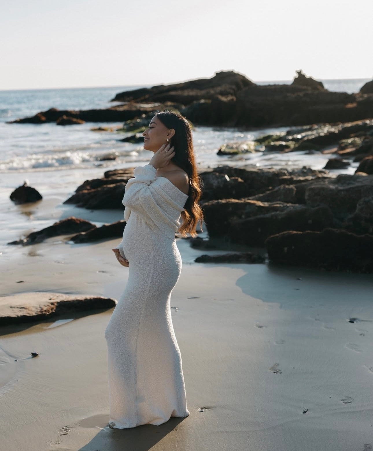 Pregnant lady standing amongst the rocks at the beach capturing her pregnancy wearing Everything Lace Hire Meshki Marisol Dress