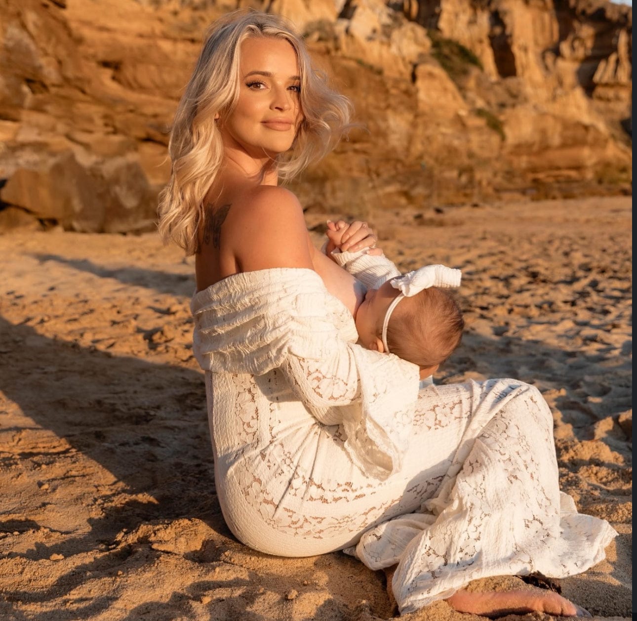 Woman in Everything Lace Hire House of CB Belle dress holding a baby on a rocky beach at sunset during a photoshoot.