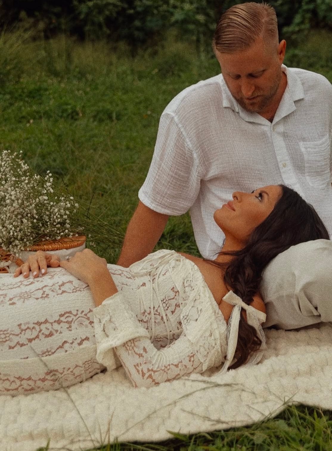 A pregnant lady announcing her pregnancy wearing Everything Lace Hire House of CB Belle Dress in ivory while lying on her partners lap.