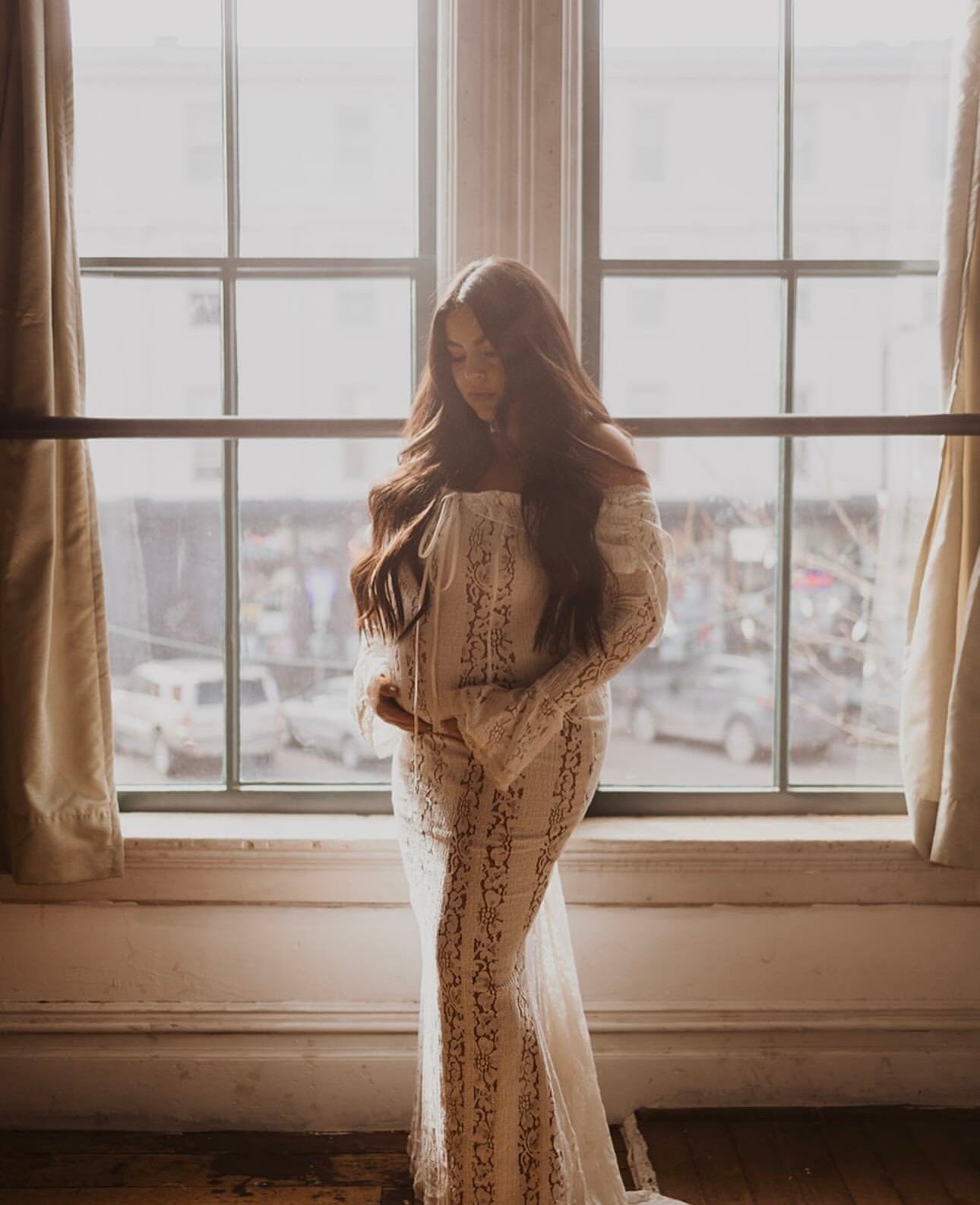 A pregnant lady holding her bump against a window back drop wearing Everything Lace Hire House of CB Belle Dress for her maternity photoshoot.