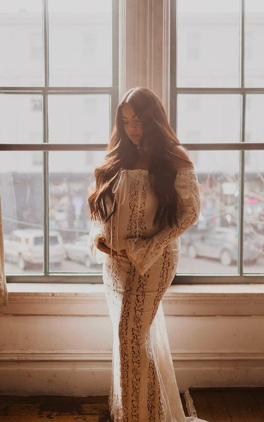 A woman standing by a large window, wearing Everything Lace Hire House of CB Belle Dress a floral lace maxi dress with a fluted hem and off-the-shoulder neckline.