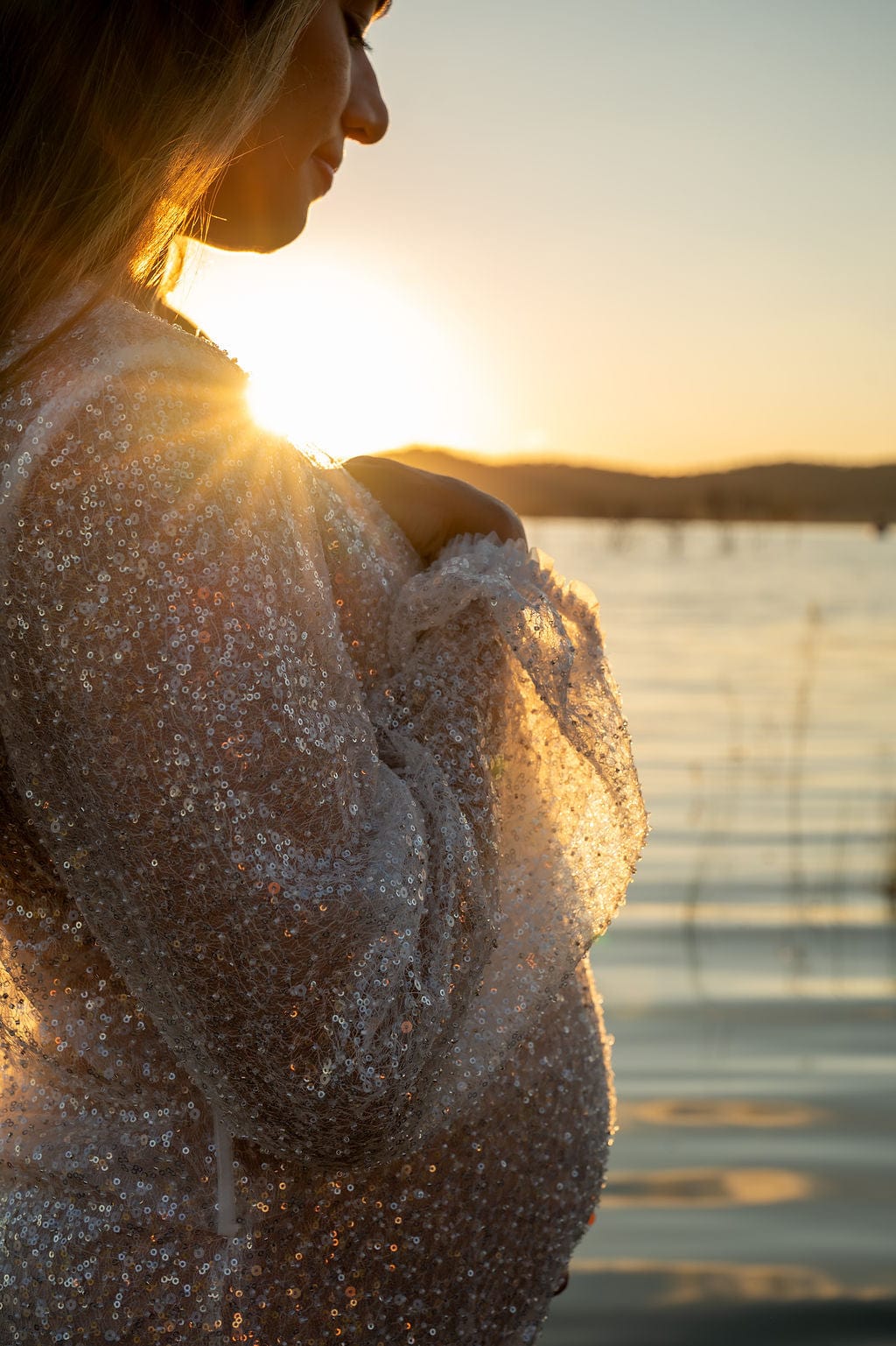 Person wearing a sequin beaded robe from Everything Lace Hire with a sunset or sunrise over water in the background during her maternity photoshoot.