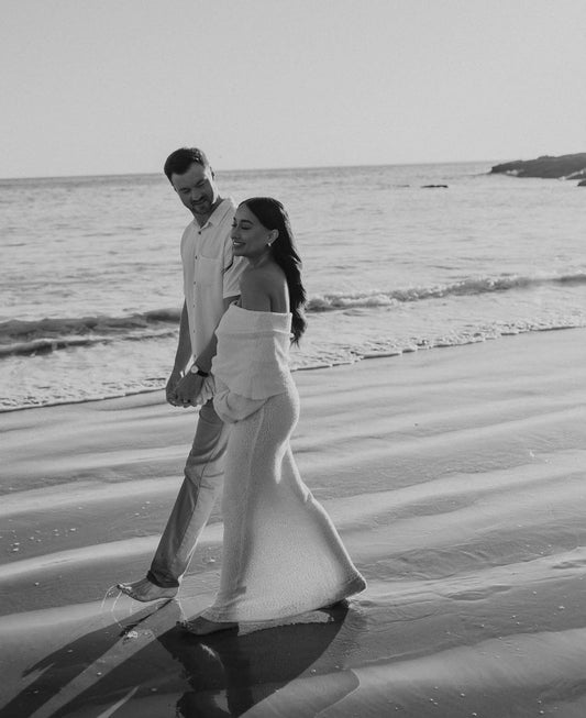 pregnant couple walking along the beach smiling and looking at each other. Pregnant mother is wearing Everything Lace Hire Meshki Marisol Dress