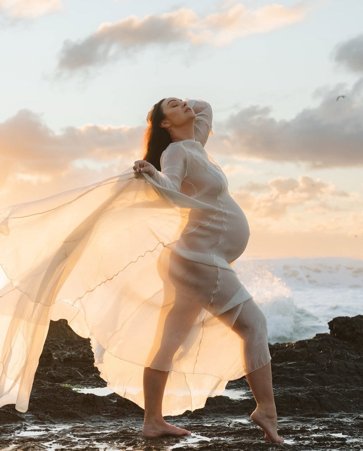 Mother standing on rocks at the beach showcasing her pregnancy wearing Everything Lace Hire Vienna Dress