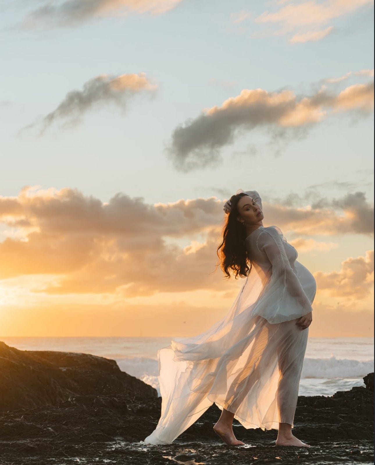 Mother standing on rocks at the beach showcasing her pregnancy wearing Everything Lace Hire Vienna Dress