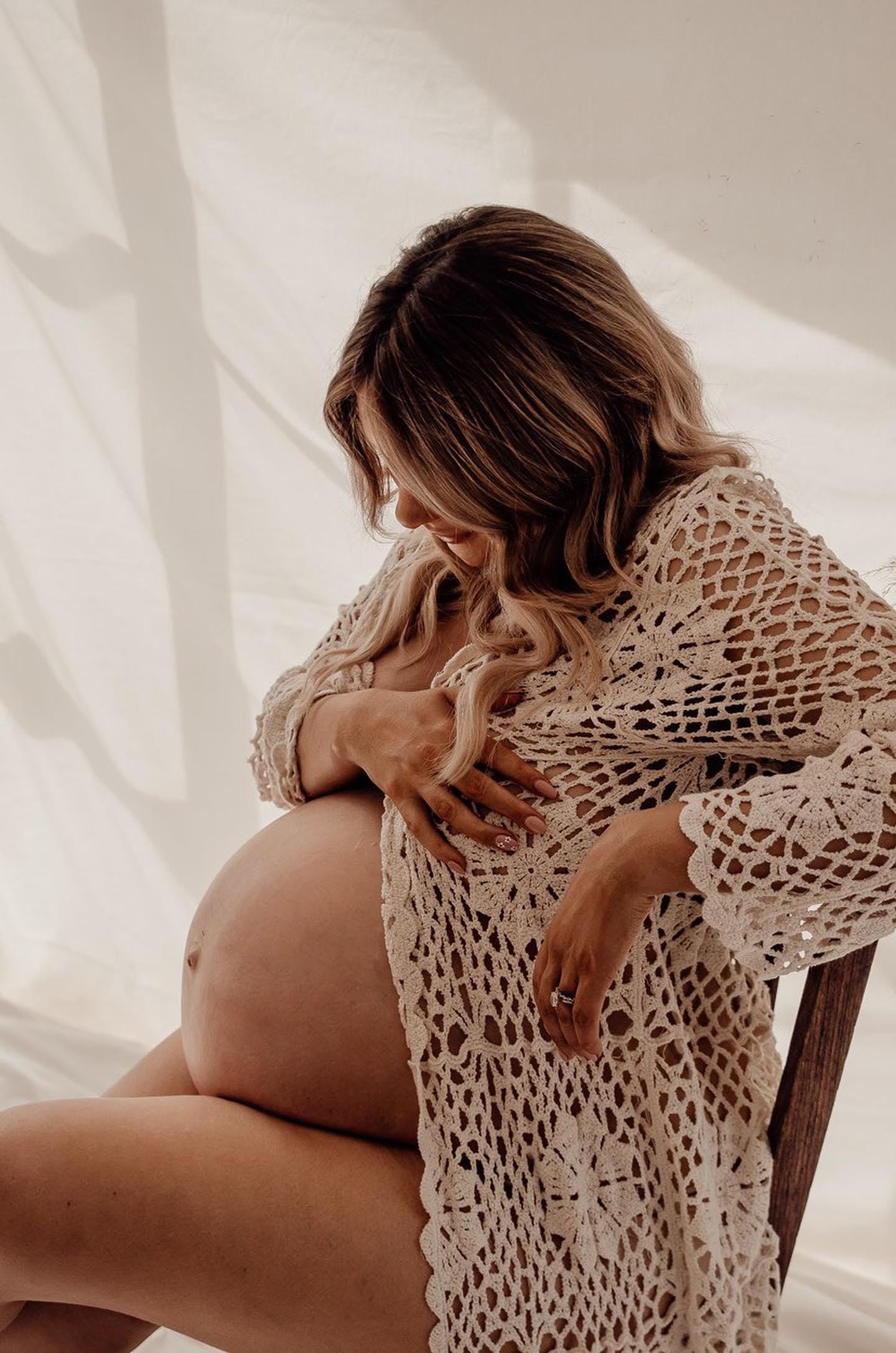 A pregnant lady its on a chair looking down at her bump during her maternity photoshoot wearing Everything Lace Hire Belle Crochet Photoshoot Robe