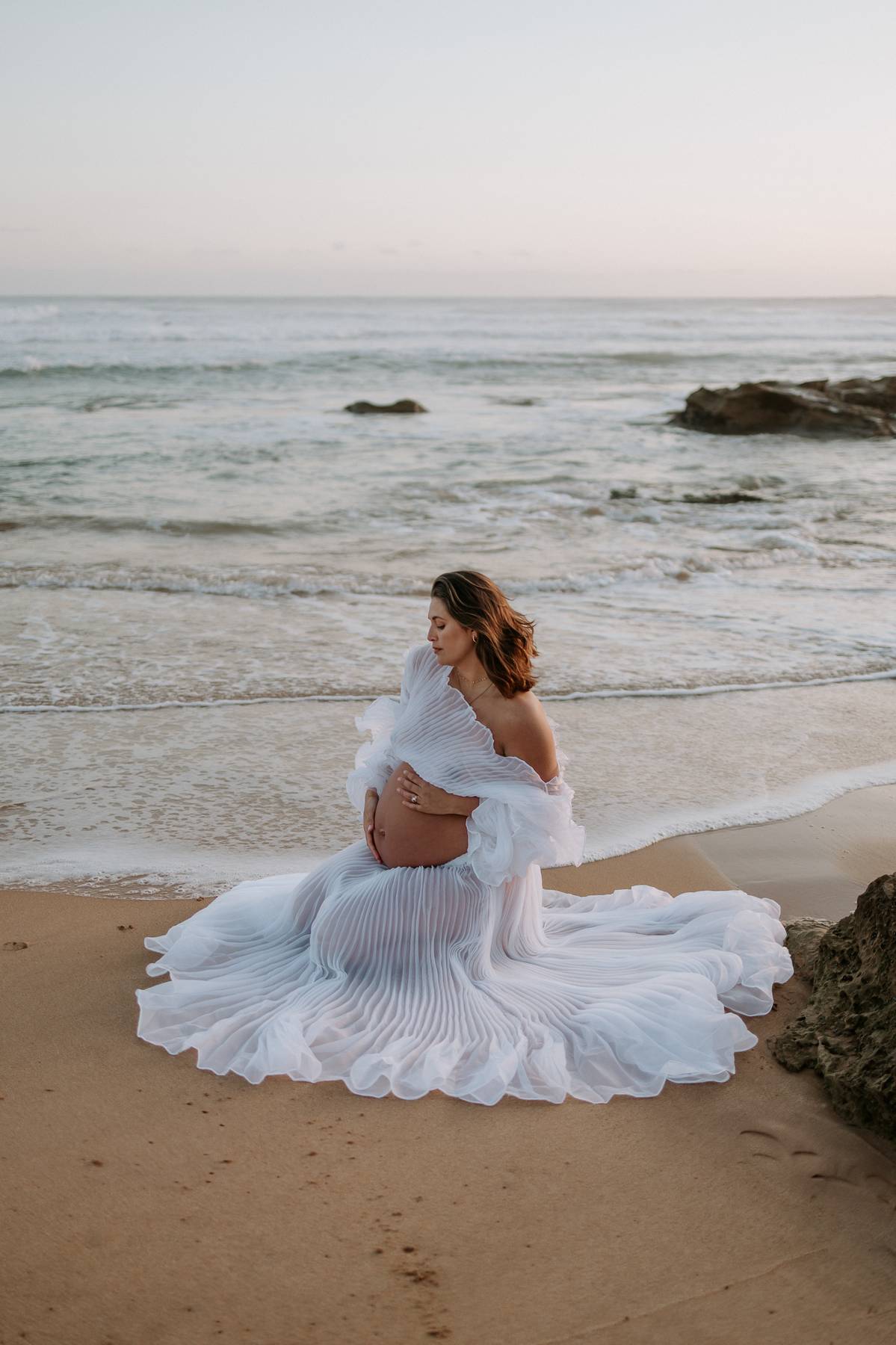 Pregnant woman wearing Everything Lace Hire Bronte Maternity Photoshoot Set dress sitting on a beach with ocean waves in the background