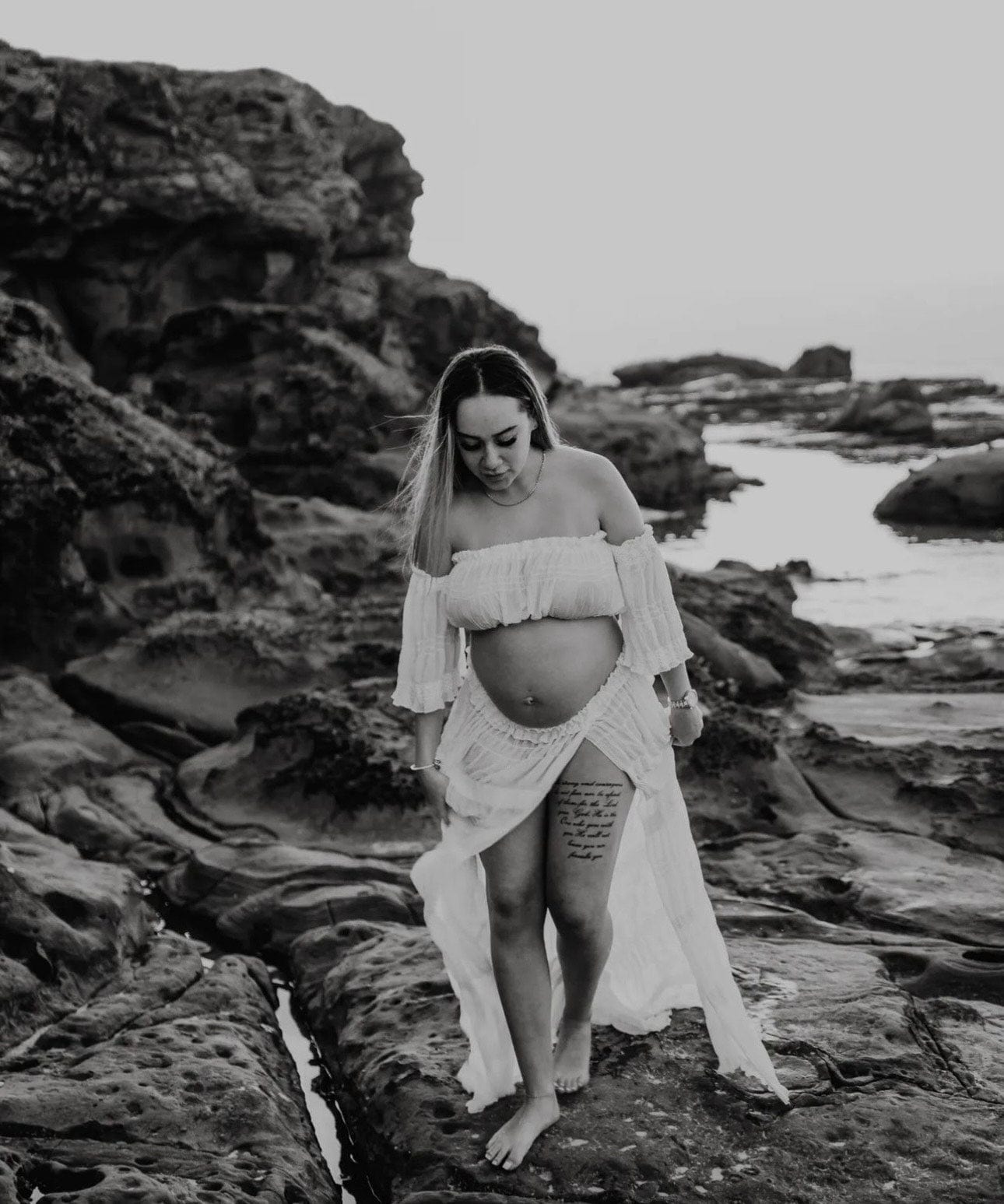 A pregnant lady walks along the rocks at the beach wearing Everything lace Hire Anabelle Two Piece Maternity Photoshoot Set