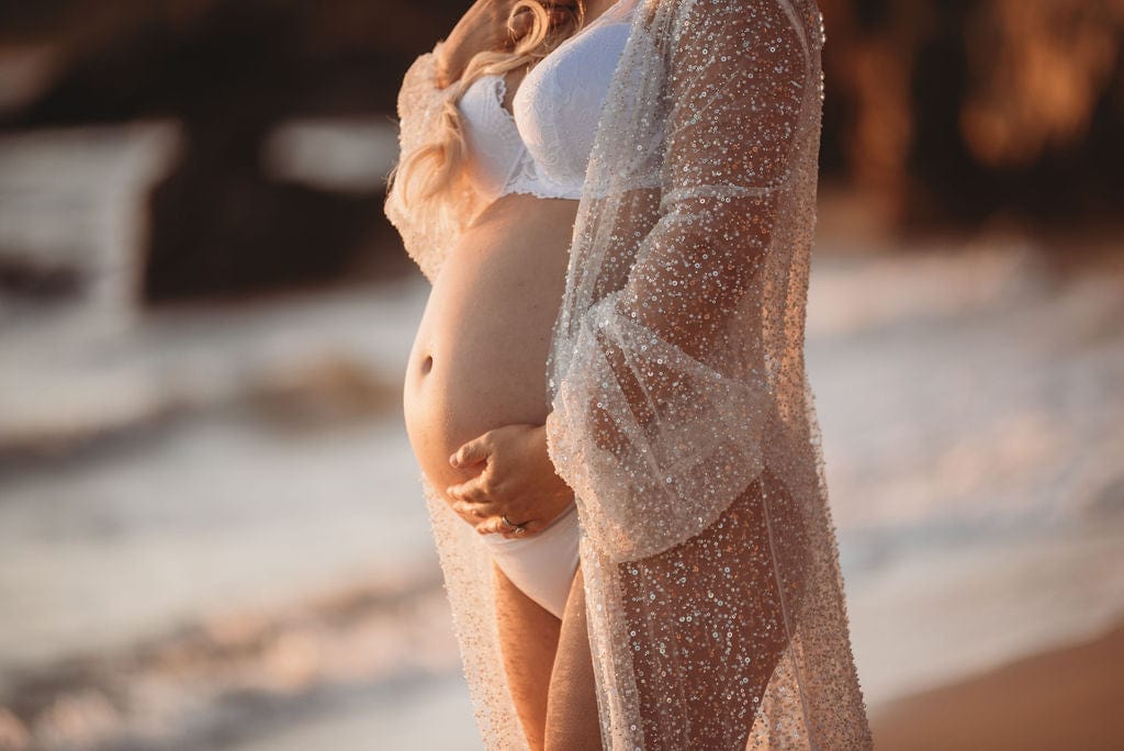 A pregnant lady stands amongst the sand on the beach holding her bump wearing Everything Lace Hire Athena Sparkly beaded maternity photoshoot robe