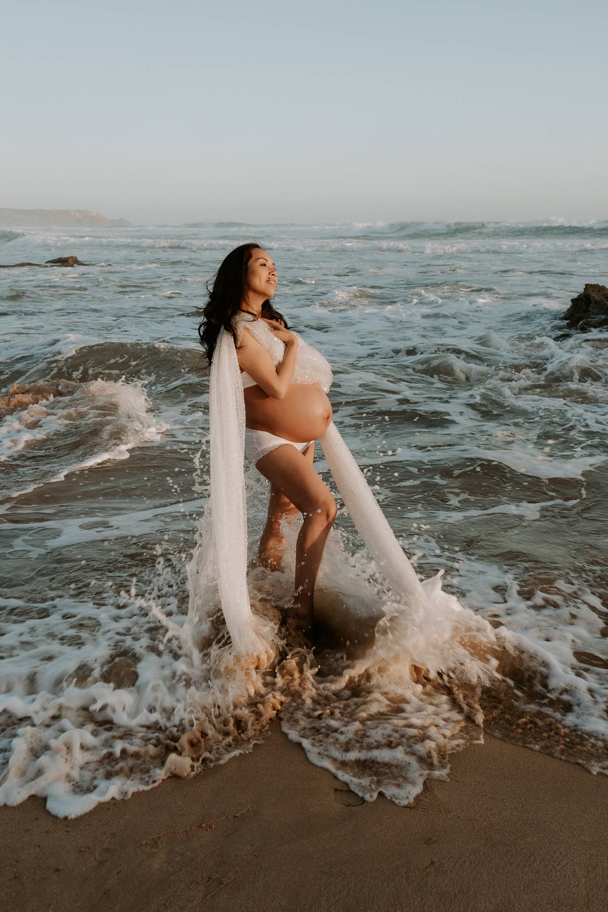Pregnant woman standing in the  ocean waves during her maternity photoshoot wearing Everything Lace Hire maternity sequin beaded drape dress over her maternity bump.
