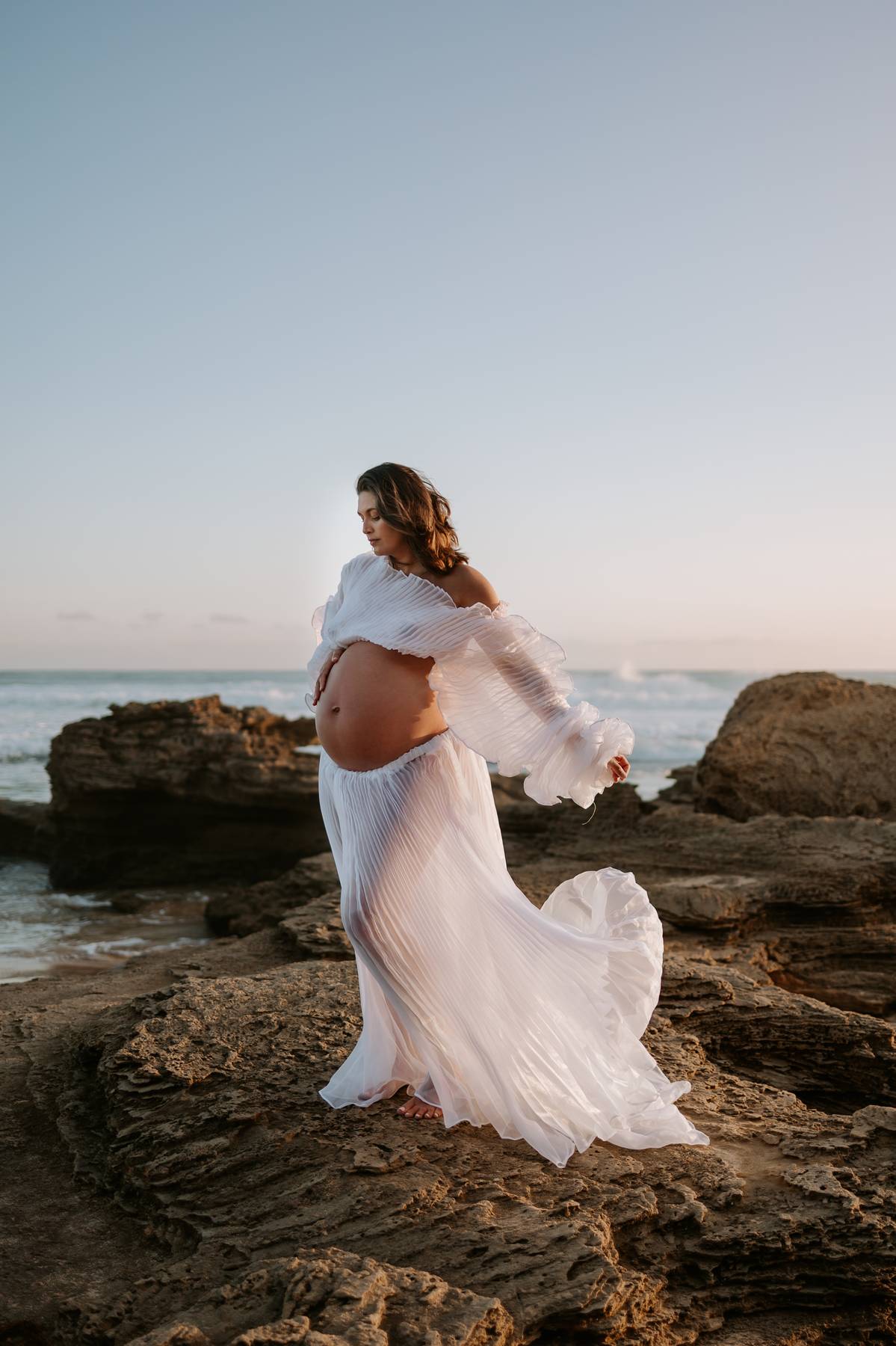 Pregnant woman in a flowing white maternity Crinkle dress from Everything Lace Hire standing on rocky beach with ocean view