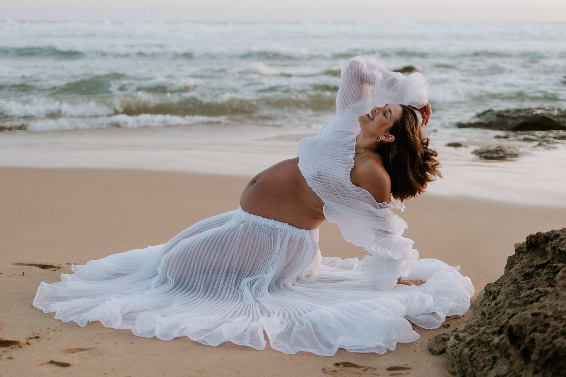 Woman Wearing Everything Lace Hire Bronte Maternity Photoshoot Set in a  sitting on a beach ocean waves in the background