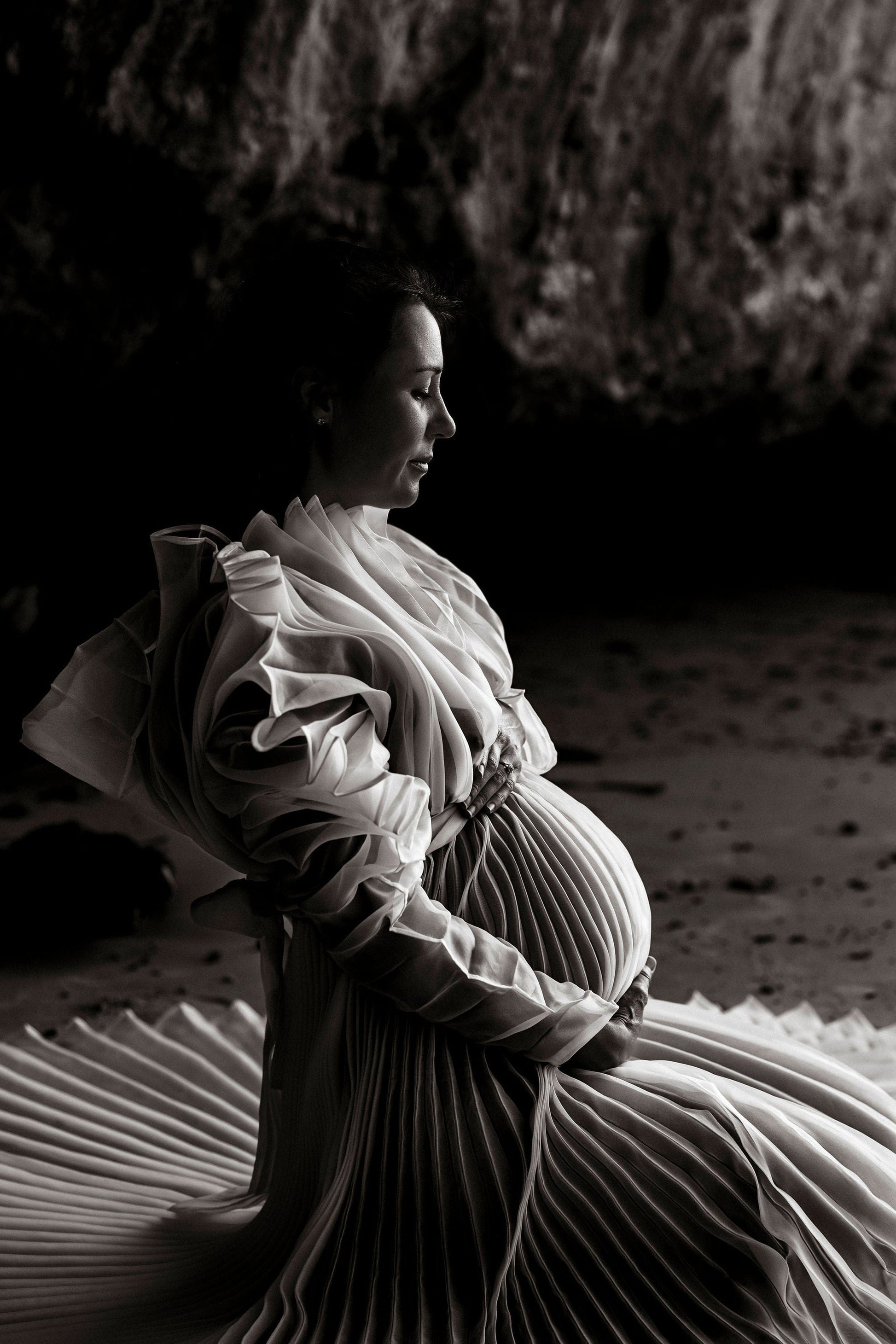 Pregnant woman in the Camille Dress a flowing pleated dress from Everything Lace Hire sitting on a sandy beach