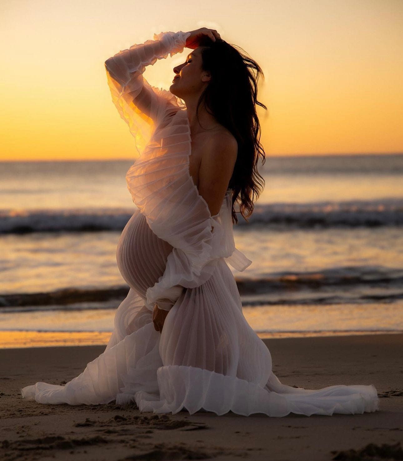 Maternity Photoshoot on the beach wearing Everything Lace Hire Camille Dress. Kneeling down, cupping her bump and holding her head