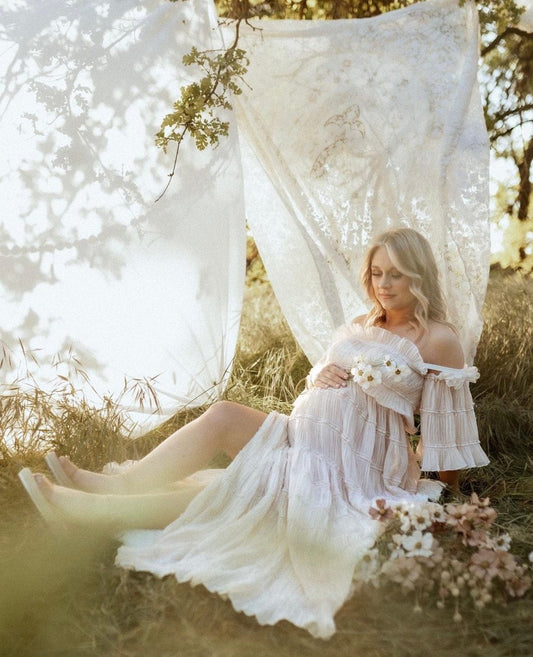 A pregnant lady sits in a fields of long grass with lace blankets hanging wearing Everything Lace Hire Chiffon Dreamer Maternity Photoshoot Dress