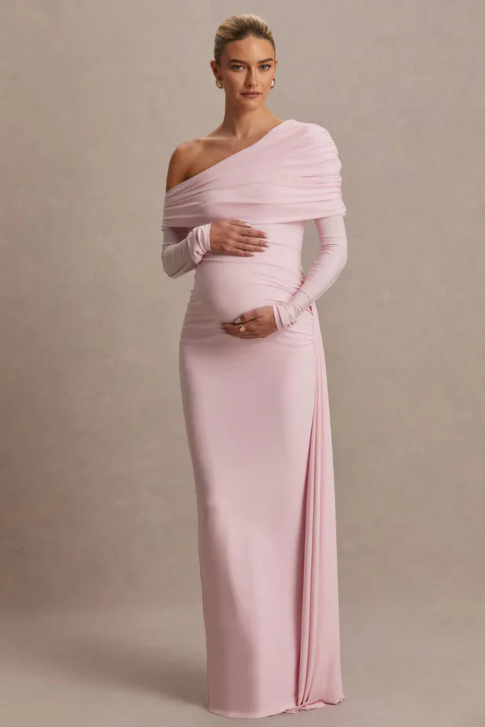 A pregnant model wearing a light pink, one-shoulder maternity gown with long sleeves and a fitted silhouette, standing against a neutral beige studio backdrop.