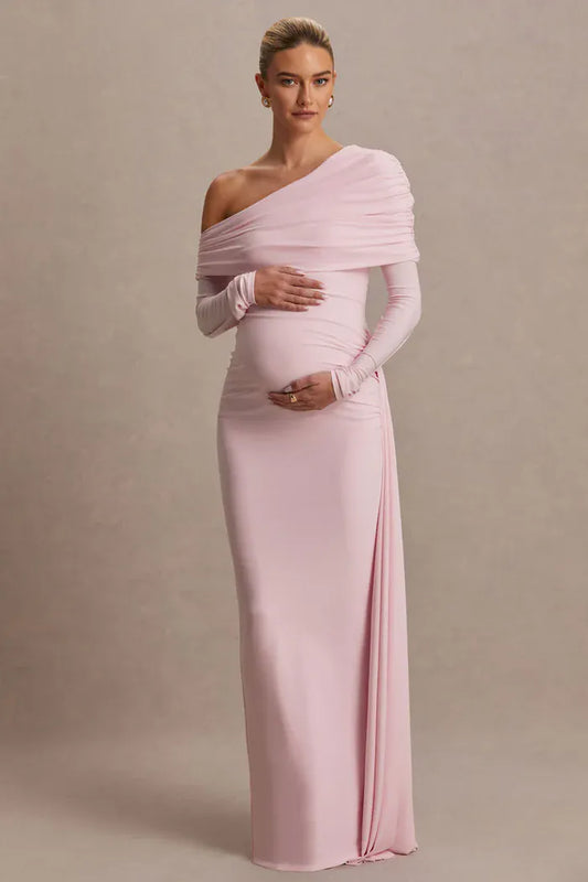 A pregnant model wearing a light pink, one-shoulder maternity gown with long sleeves and a fitted silhouette, standing against a neutral beige studio backdrop.