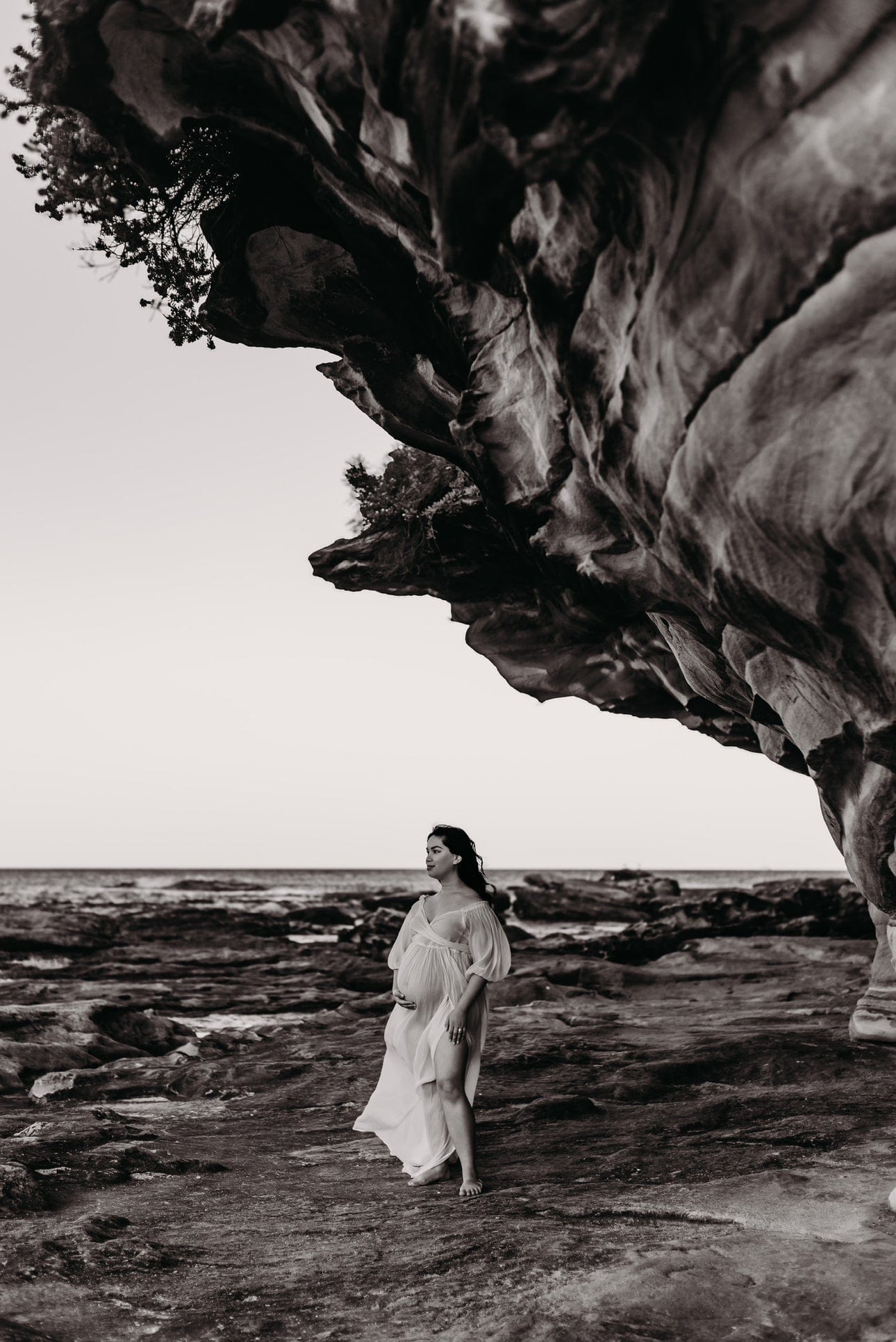 A pregnant lady looking out at sea standing on the rock edge wearing a dress from Everything Lace Hire Co & Ry Illoura Gown