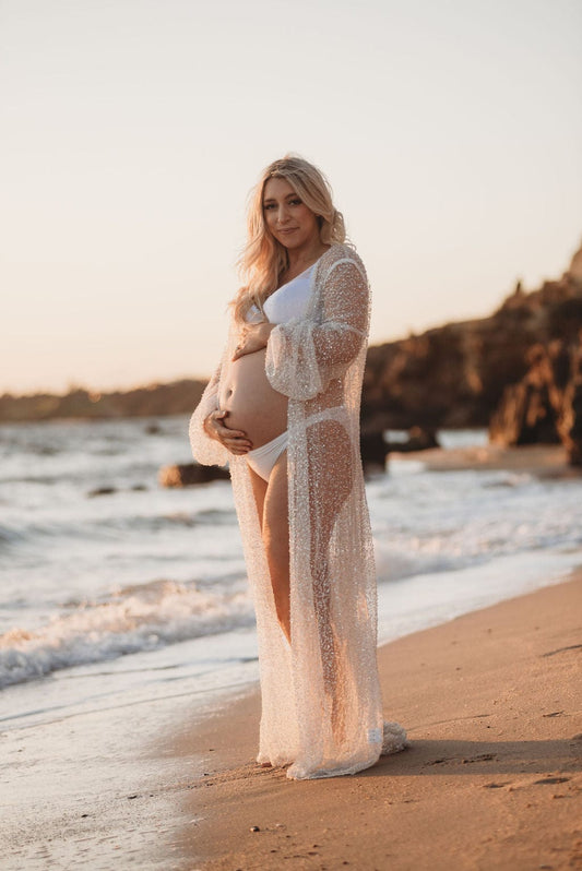 A beautiful pregnant lady stands on the beach holding her bump during her maternity photoshoot wearing Everything Lace Hire's Homeboddi Athena Beaded Robe. 