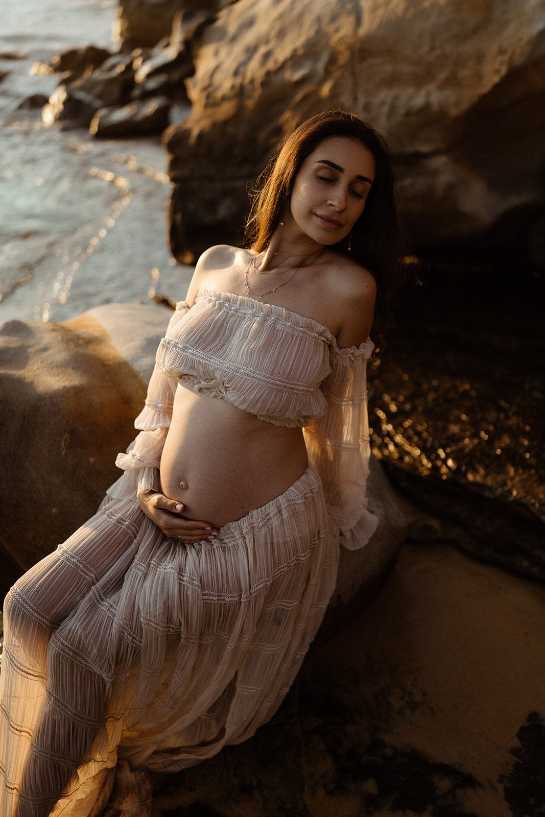 Pregnant woman in a flowing maternity outfit from Everything Lace Hire sitting on rocks by the water