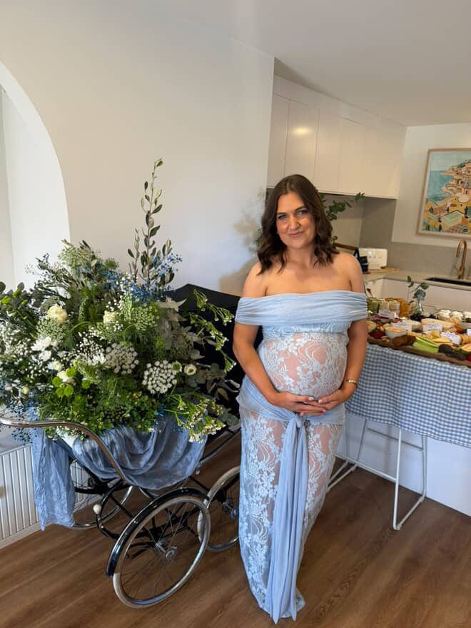 A pregnant lady wearing Everything Lace Hire Gracie Twist Maxi Baby Shower Dress in a light blue standing at her baby shower with flowers and a table.