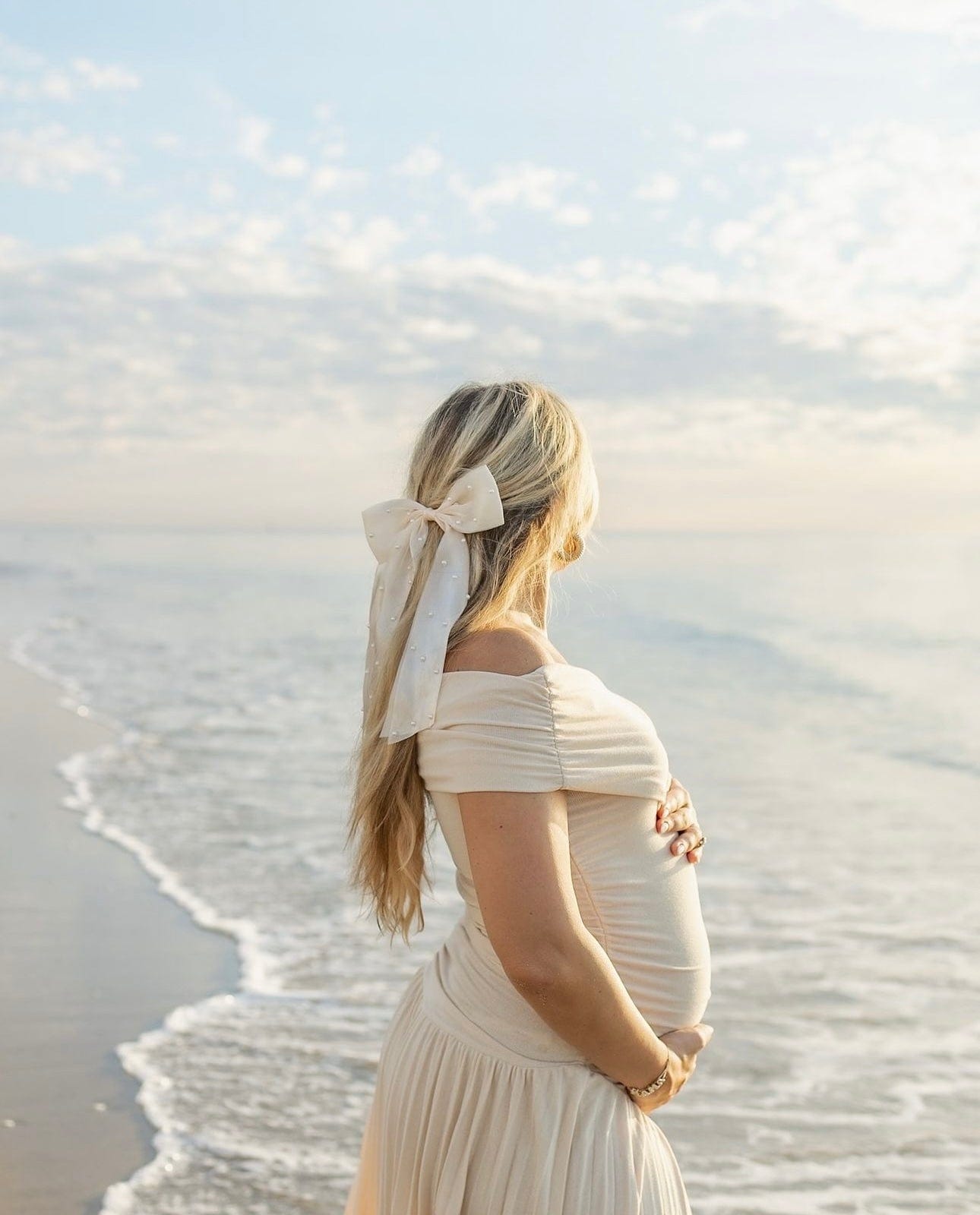 A pregnant lady stands in the ocean looking out to sea wearing Everything Lace Hire Field Of Dreams Baby Shower Dress