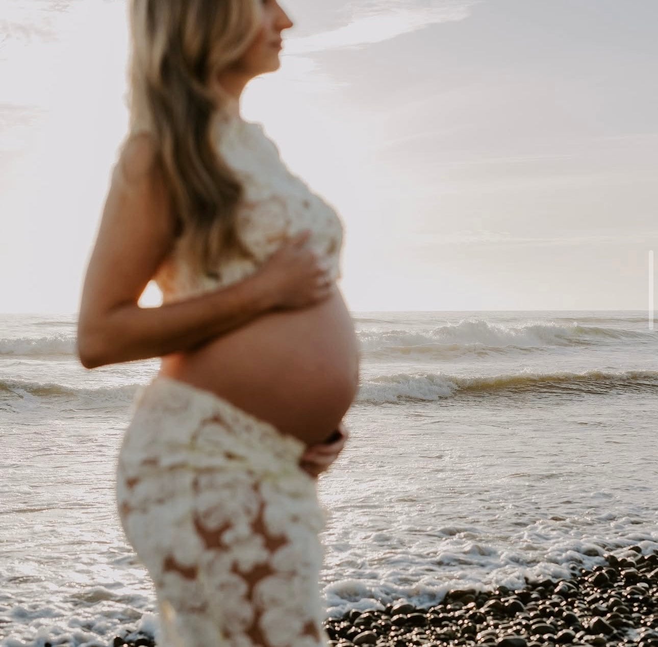 Pregnant woman in Everything Lace Hire Fiorella Set showcasing her pregnancy bump standing on a beach with waves in the background