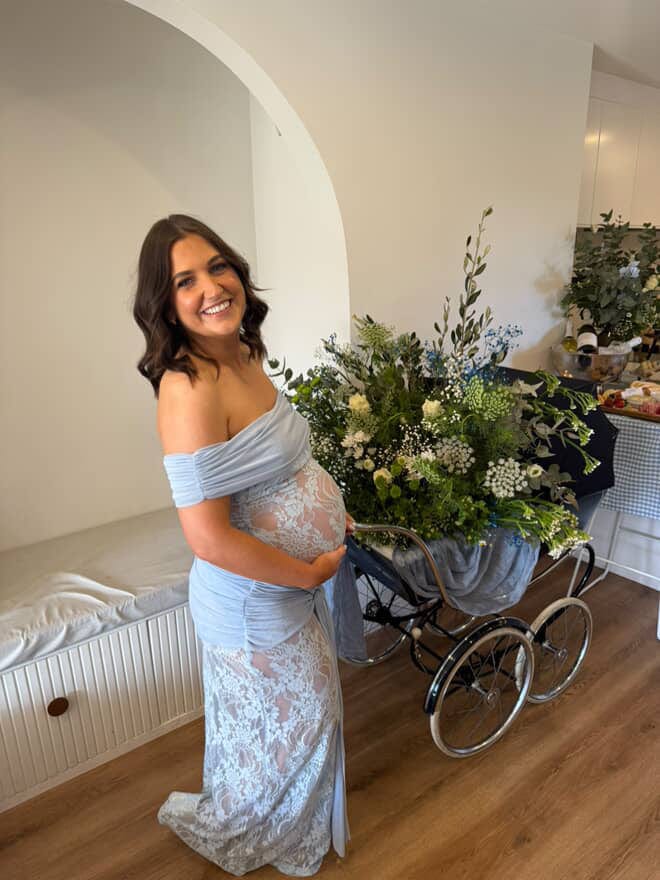 Pregnant lady in a light blue off-shoulder dress baby shower dress from Everything Lace Hire standing next to a baby carriage with flowers at her baby shower.