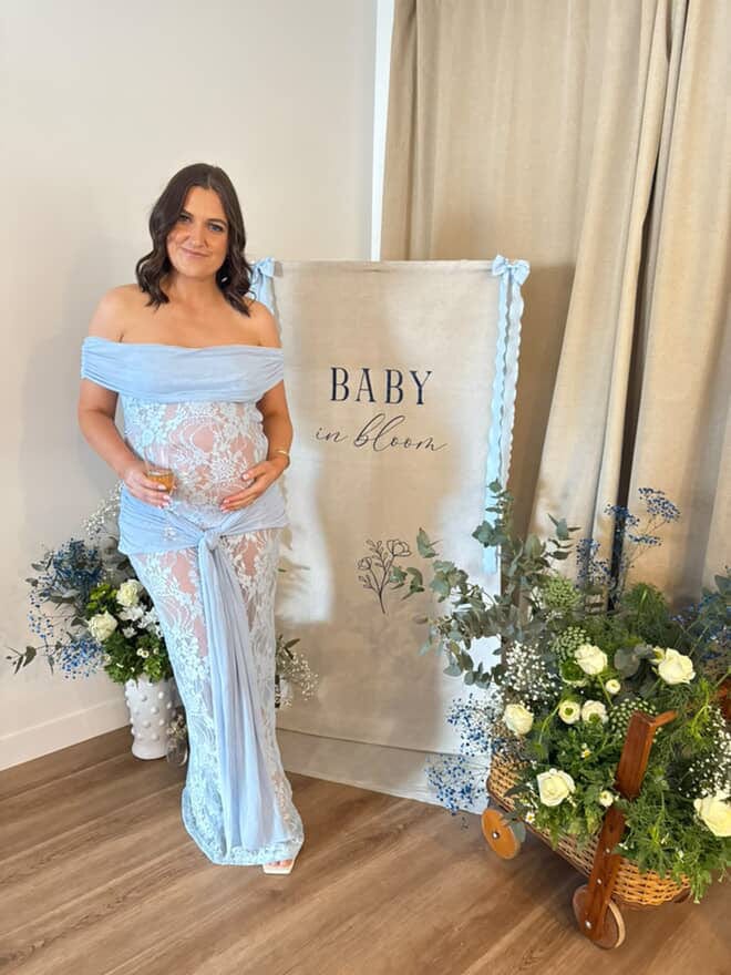 Pregnant woman in a light blue lace dress from Everything Lace Hire standing next to a 'Baby in Bloom' sign with floral decorations during her baby shower.
