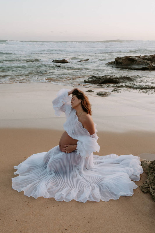 Pregnant woman in a flowing crinkle white two piece dress from Everything Lace Hire on a beach during her maternity photoshoot