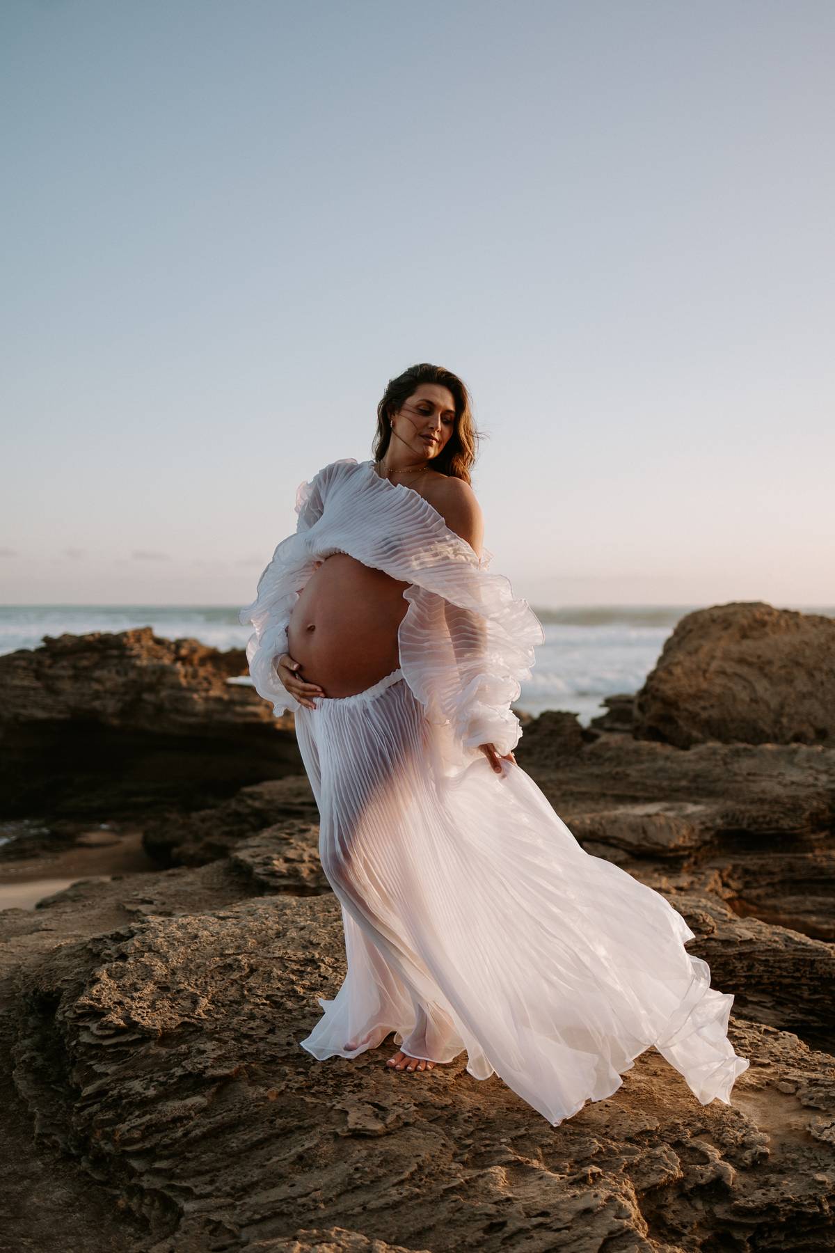 Pregnant woman in a flowing white crinkle two piece set dress from Everything Lace Hire standing on rocky beach at sunset.