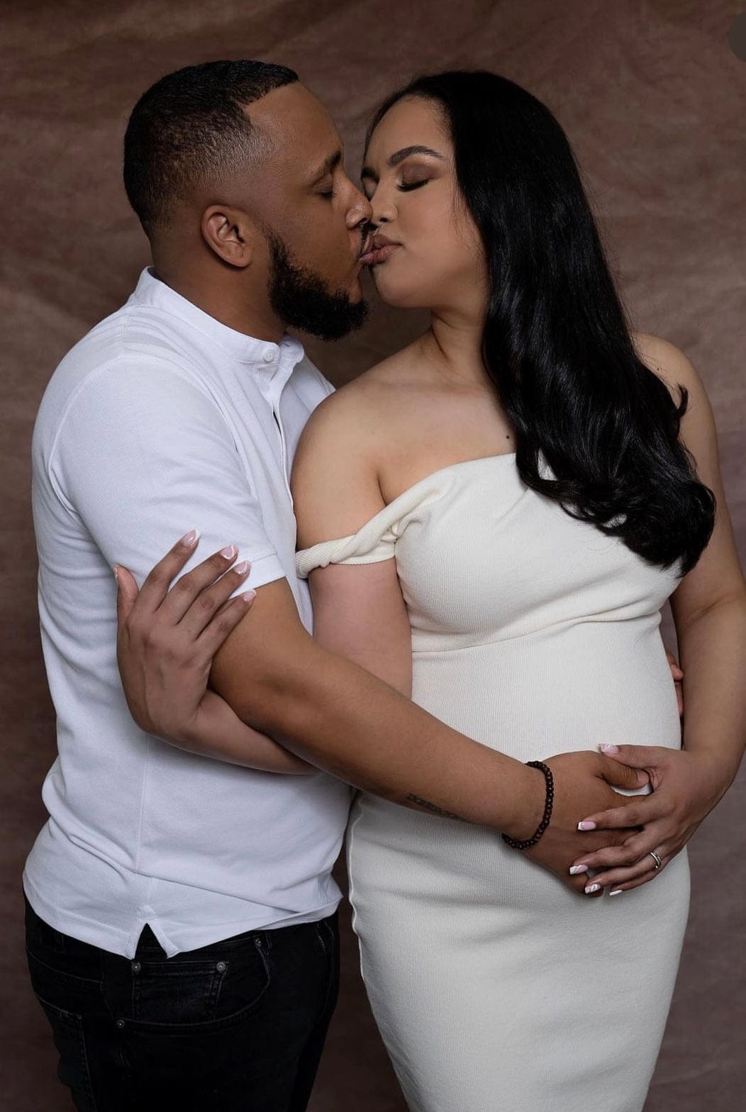 A couple stand in each others arms kissing celebrating her pregnancy wearing Everything Lace Hire Johansen Poppy Dress in white a fitted maternity dress