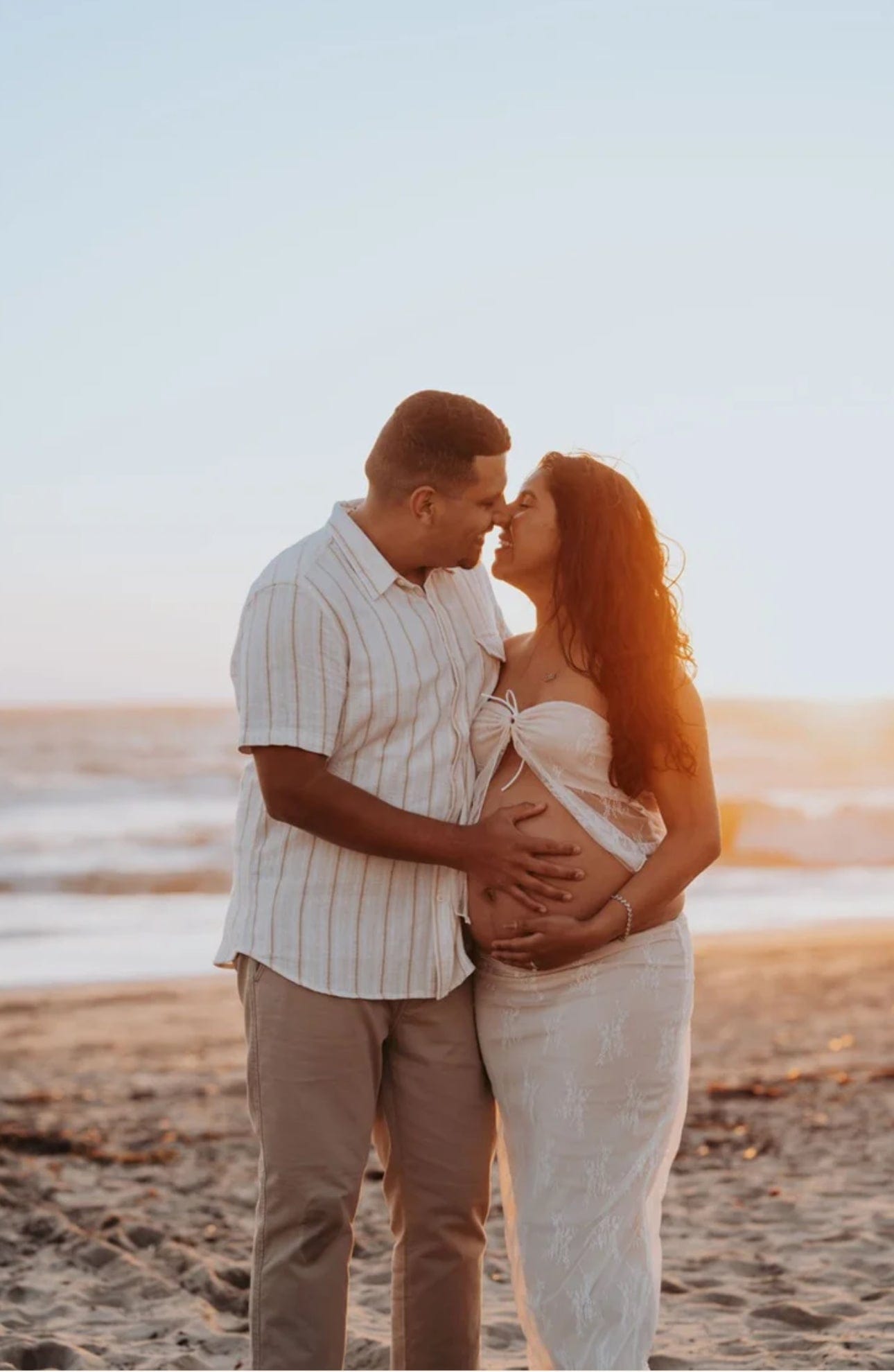 Man and woman standing on a beach, man holding woman's pregnant belly wearing Everything Lace Hire Karolina Lace Set.