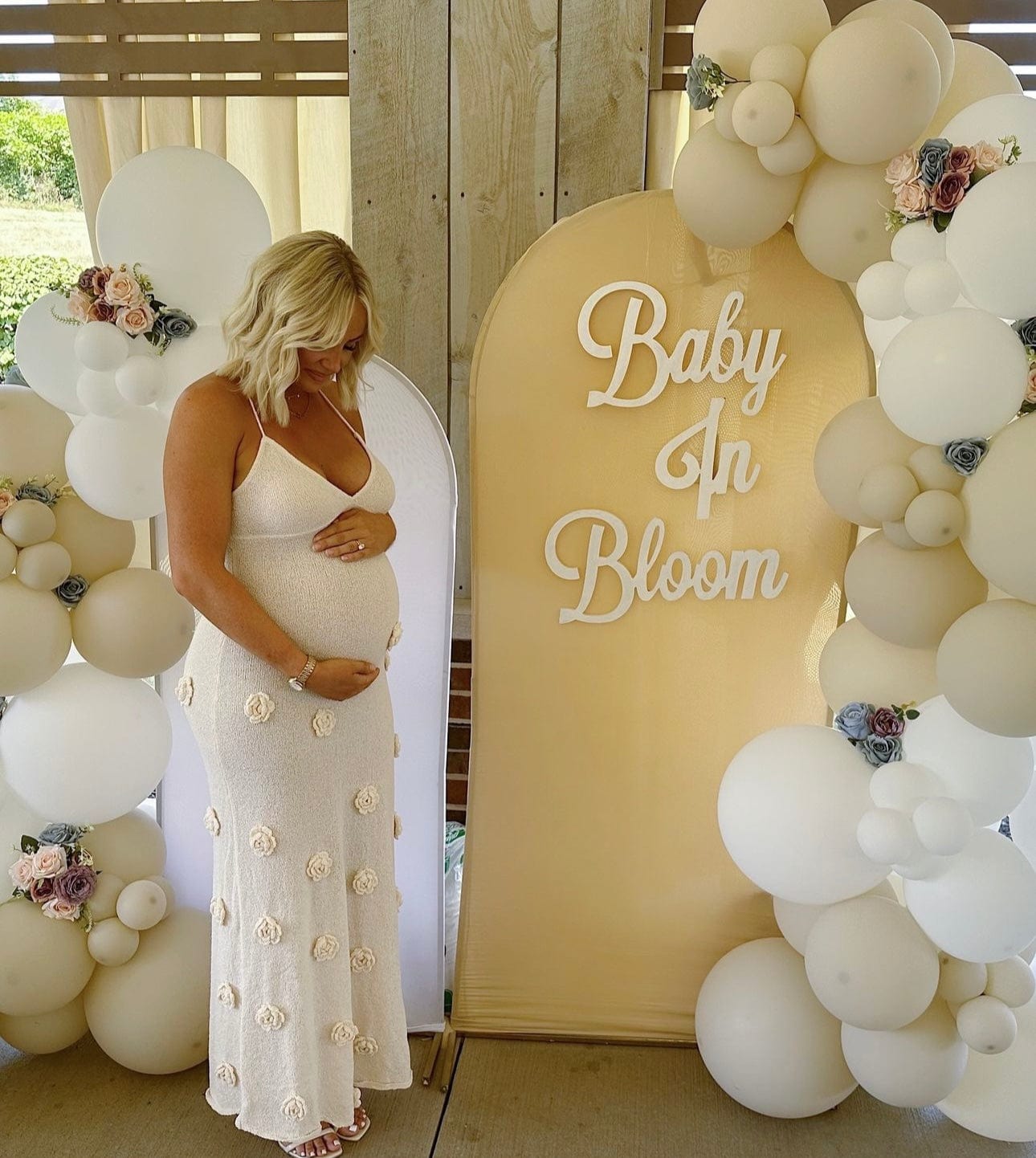 A pregnant lady looking down at her bump during her baby shower wearing Everything Lace Hire Meshki Suki Blossom Baby Shower Dress a knitted piece with delicate florals throughout