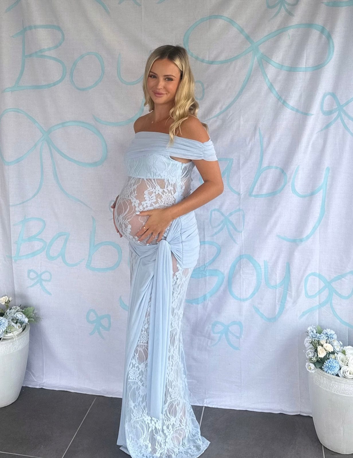 Pregnant woman wearing Everything Lace Hire Gracie Twist a lace maternity Baby Shower dress standing in front of a Baby Boy Sign backdrop.