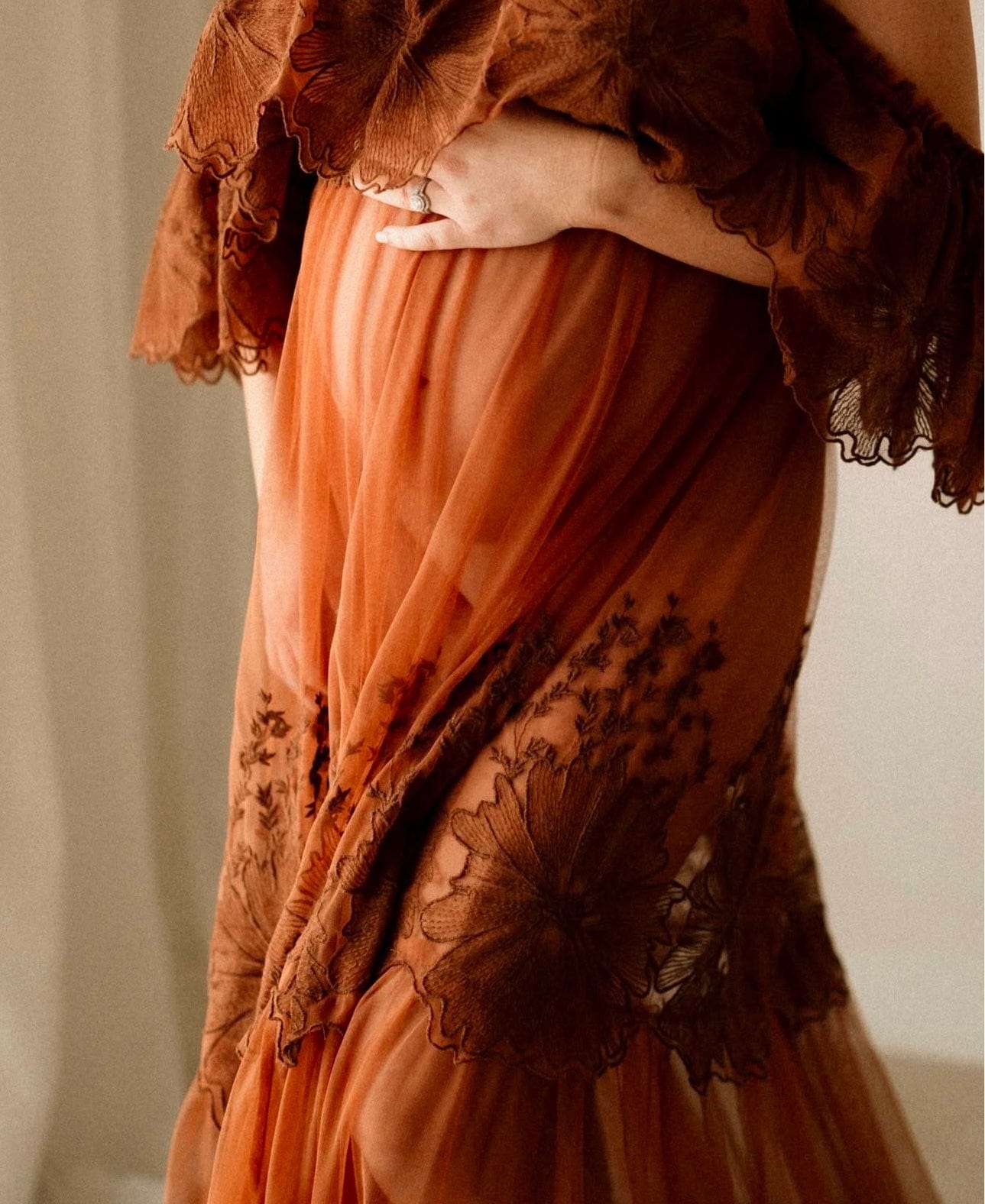 A pregnant mother holding her bump during her maternity photoshoot wearing Everything Lace Hire Euphoria Off the Shoulder Dress in rust