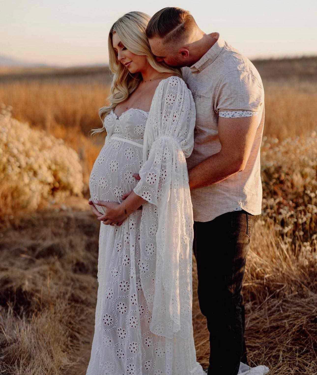 A glowing mumma celebrating her pregnancy with her partner wearing Everything Lace Hire's Gwendolyn Gown during her maternity photoshoot in a field.