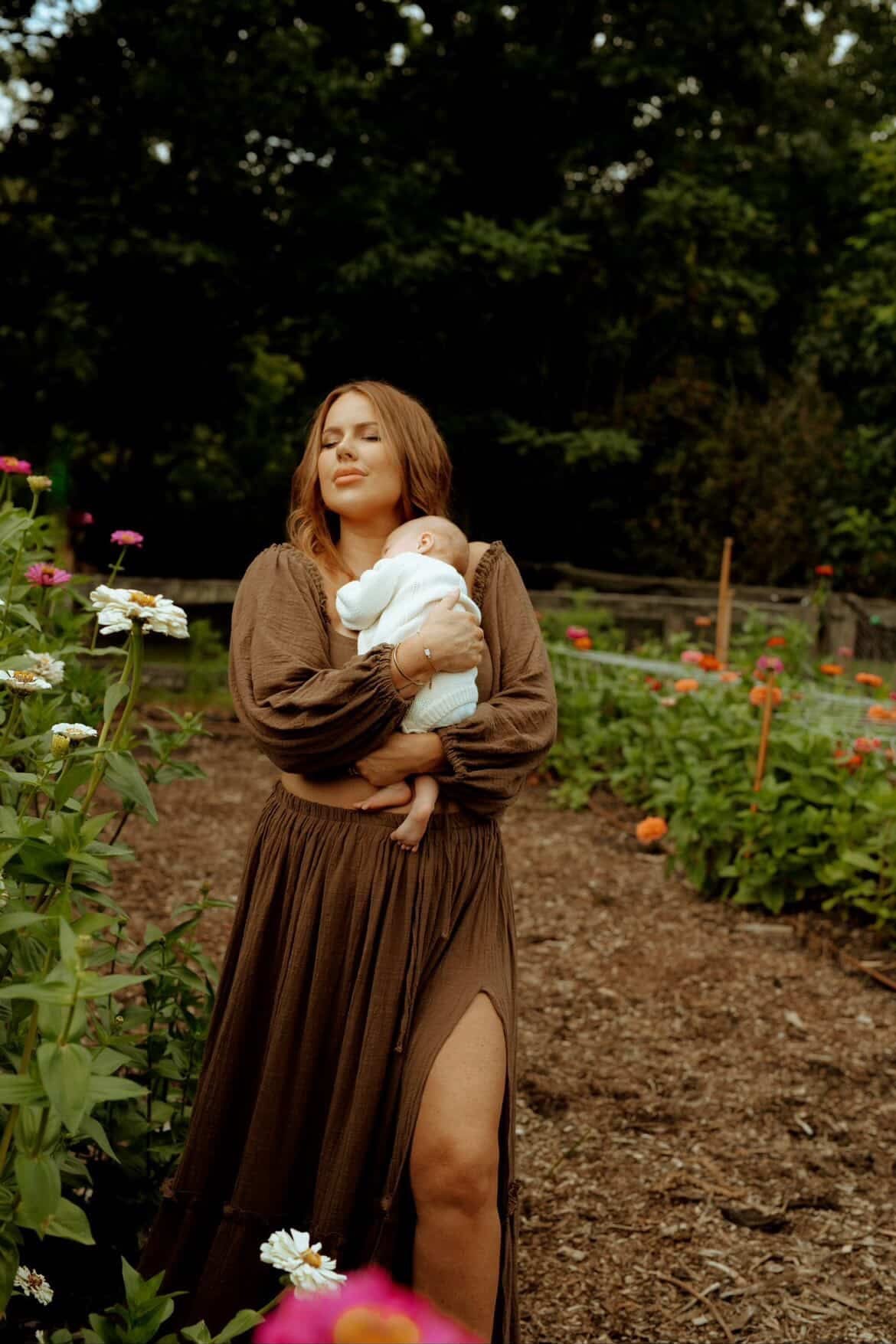 A mother holding  her baby in a garden with flowers and trees in the background wearing Everything Lace Hire chocolate brown Love Me Two Piece Set