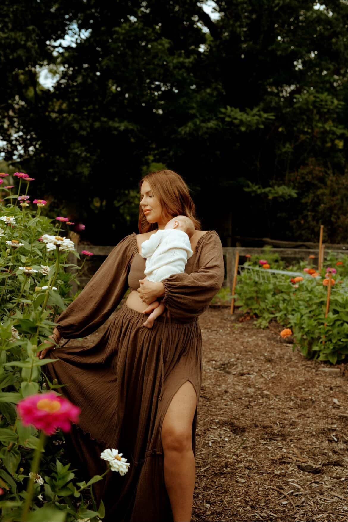 A mother holding  herbaby in a garden with flowers and trees in the background wearing Everything Lace Hire Brown Love Me Set for her motherhood photoshoot