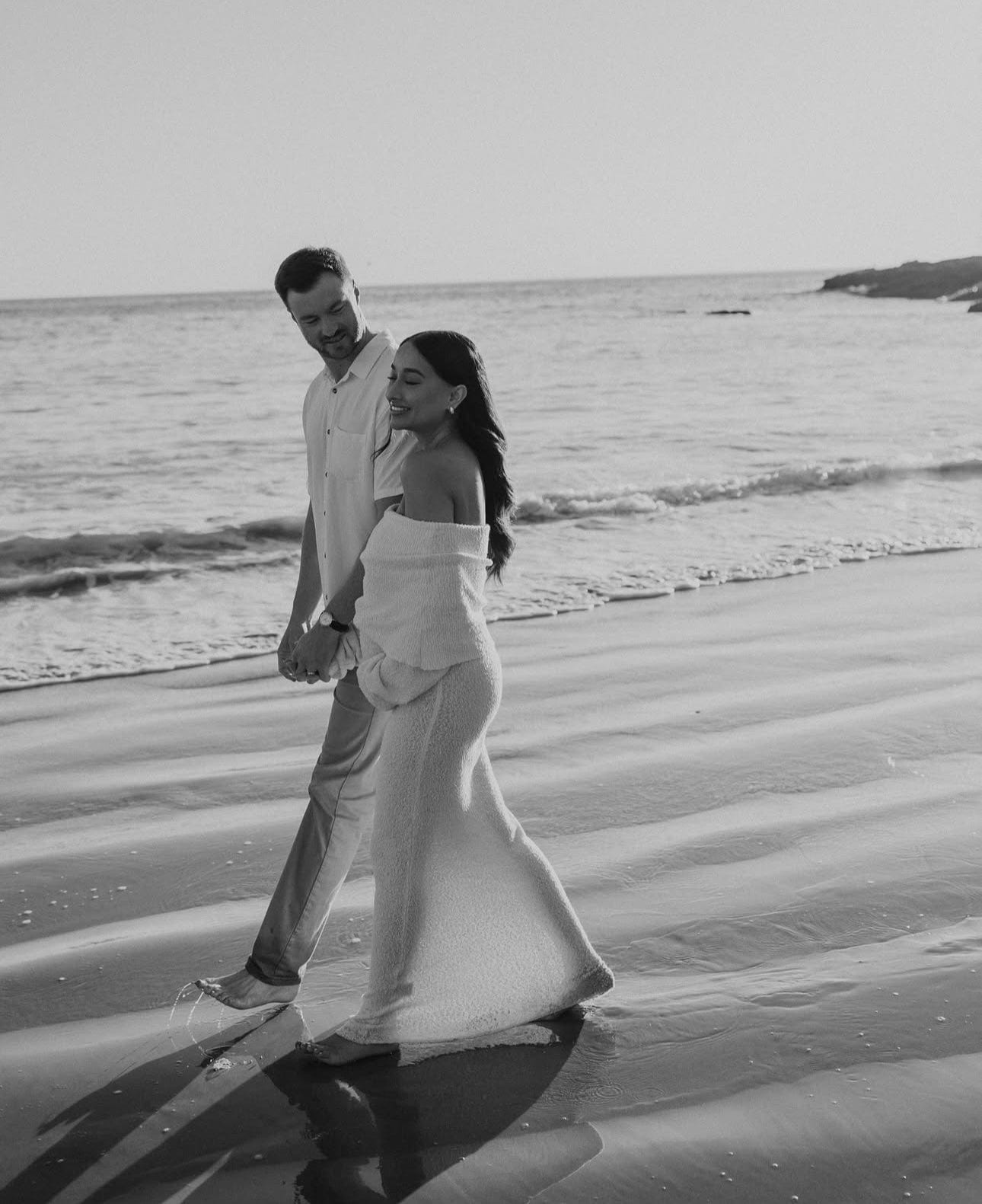 pregnant couple walking along the beach smiling and looking at each other. Pregnant mother is wearing Everything Lace Hire Meshki Marisol Dress