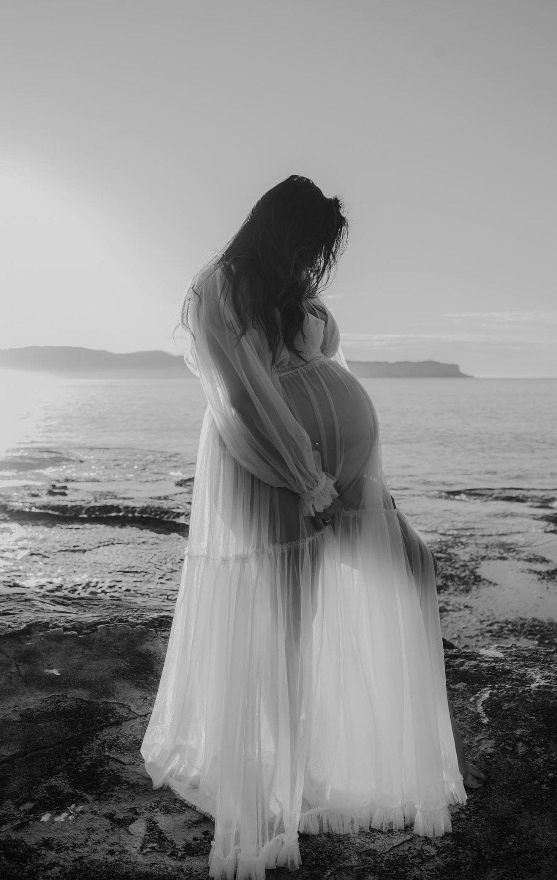 A pregnant mother standing on the rocks at the beach looking down at her bump wearing Everything Lace Hire Wish For it Gown