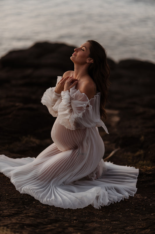 Pregnancy photoshoot on the beach wearing Everything Lace Hire Camille Dress which is a white pleated off the shoulder maternity dress. She is kneeling down on the rock arms to her belly