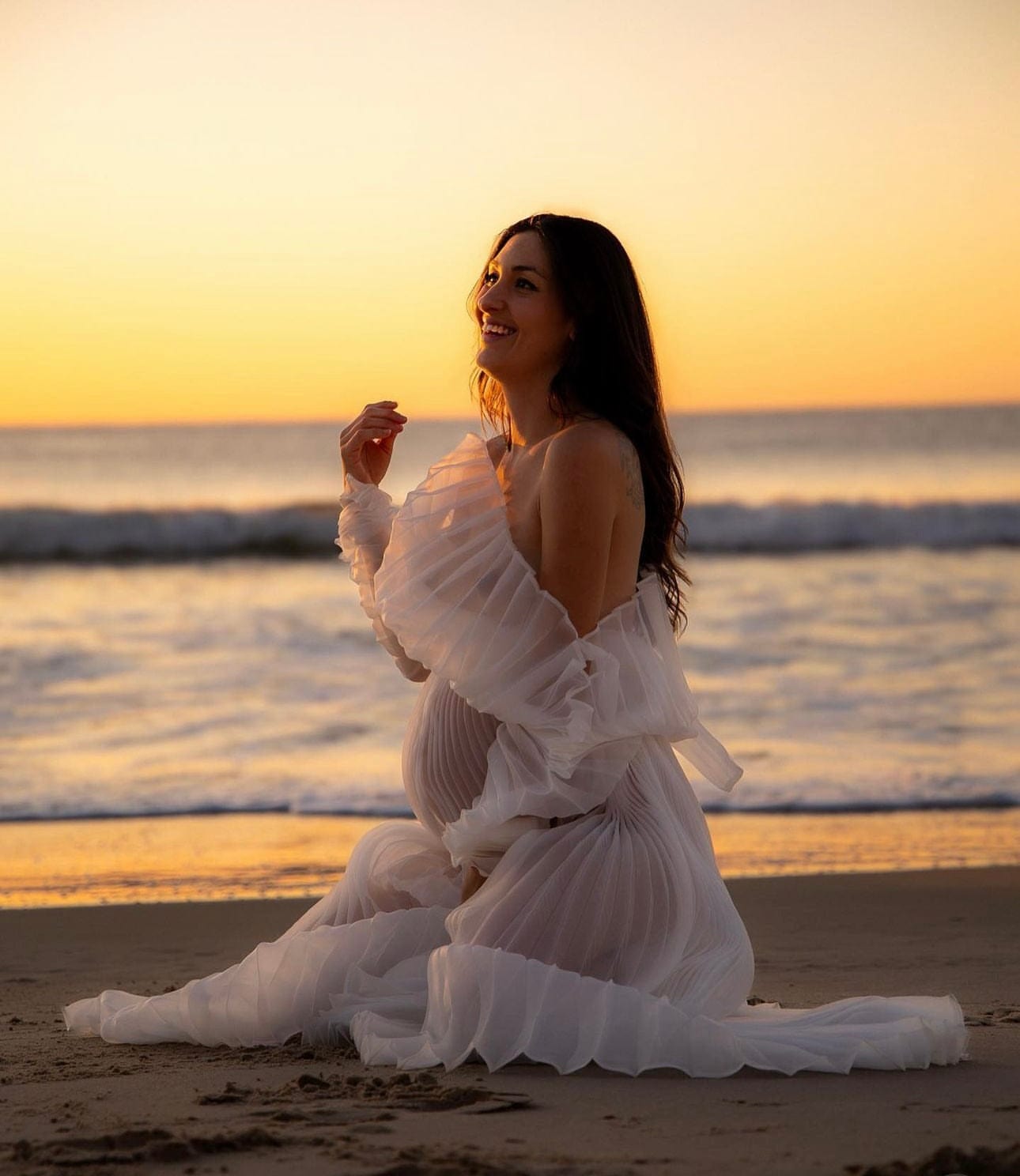 Maternity Photoshoot at the beach with mum to be kneeling down and enjoying the moments of her pregnancy wearing Everything Lace Hire Camille Maternity Photoshoot Dress