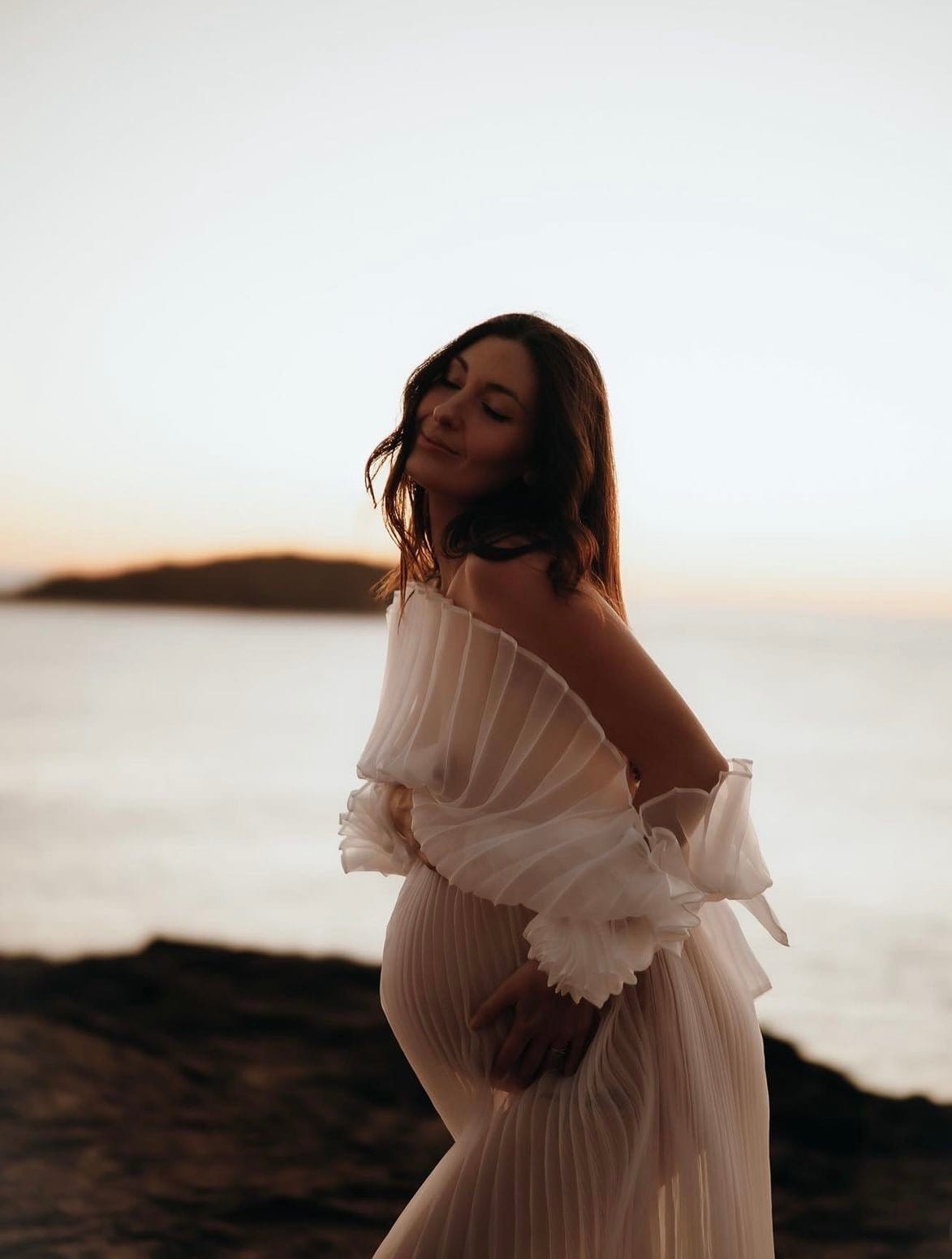 Maternity Photoshoot at the beach wearing Everything Lace Hire Camille Dress as she cups her belly on the rocks