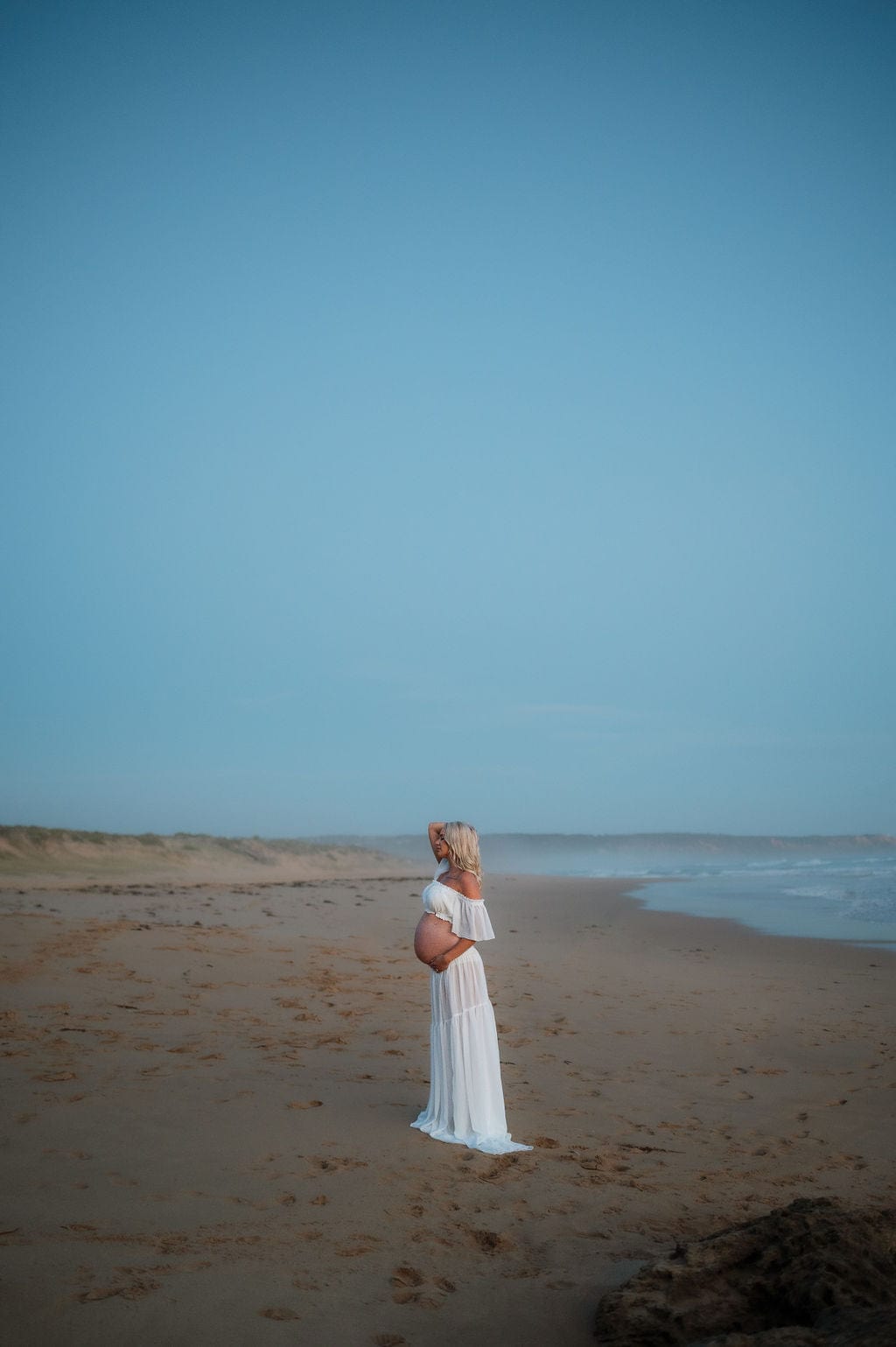 Maternity Photoshoot on Melbourne beach wearing Everything Lace Amara Photoshoot Set that is a long flowy white chiffon piece that catches the wind